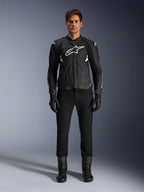 Alpinestars Dusk Airflow Leather Jacket, nero e bianco, giacca da moto in pelle con pannelli traforati per il flusso d'aria, logo Astars bianco sul petto, indossata da un pilota uomo con pantaloni neri, guanti e stivali da moto, scatto professionale in studio per equipaggiamento moto sportivo