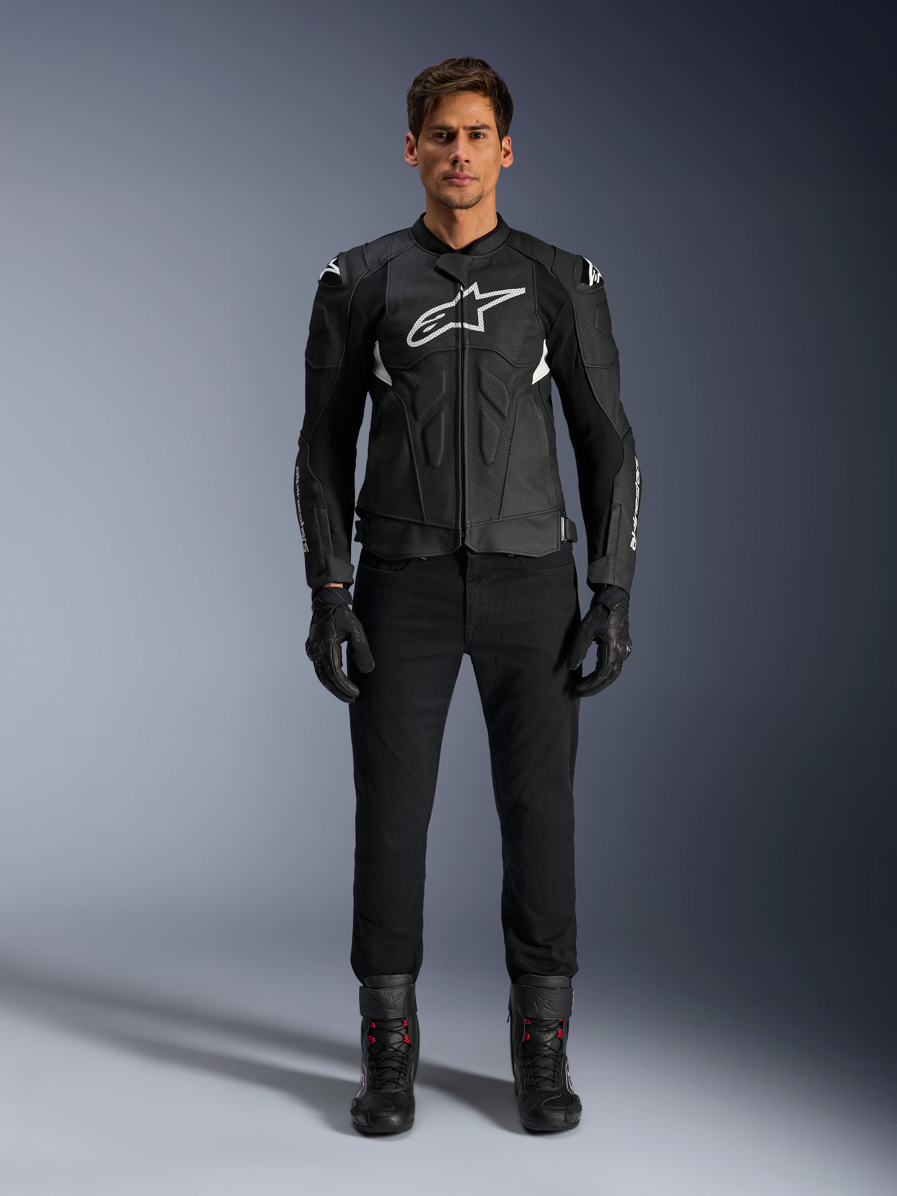 Alpinestars Dusk Airflow Leather Jacket, nero e bianco, giacca da moto in pelle con pannelli traforati per il flusso d'aria, logo Astars bianco sul petto, indossata da un pilota uomo con pantaloni neri, guanti e stivali da moto, scatto professionale in studio per equipaggiamento moto sportivo