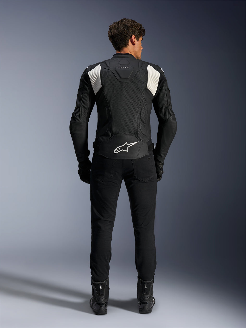 Giacca in pelle Alpinestars Dusk Airflow, nero e bianco, vista posteriore di un pilota con pantaloni e stivali neri, caratterizzata da pelle traforata per la ventilazione, pannelli a fisarmonica bianchi sulle spalle e logo bianco sulla parte bassa della schiena