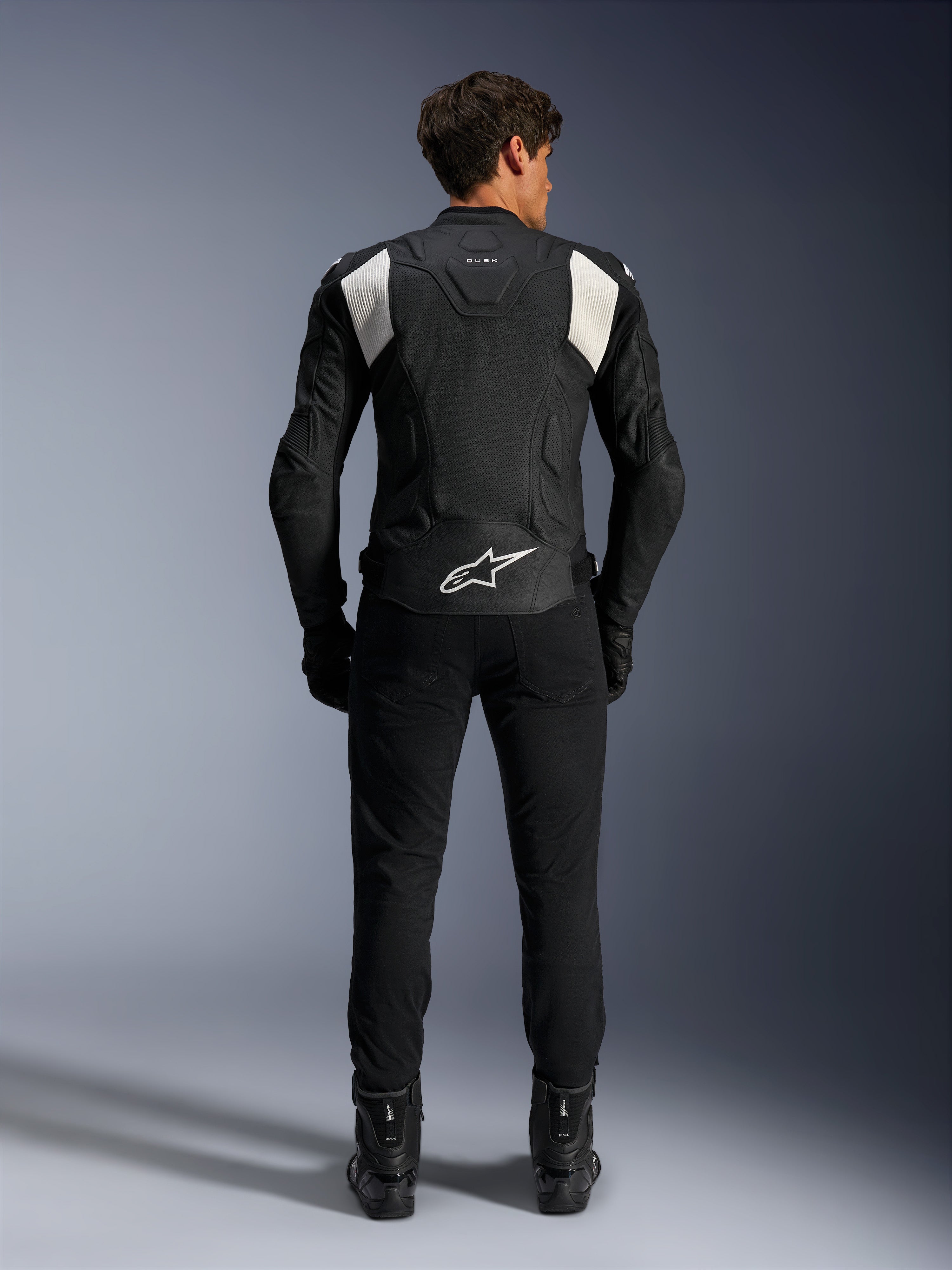 Giacca in pelle Alpinestars Dusk Airflow, nero e bianco, vista posteriore di un pilota con pantaloni e stivali neri, caratterizzata da pelle traforata per la ventilazione, pannelli a fisarmonica bianchi sulle spalle e logo bianco sulla parte bassa della schiena