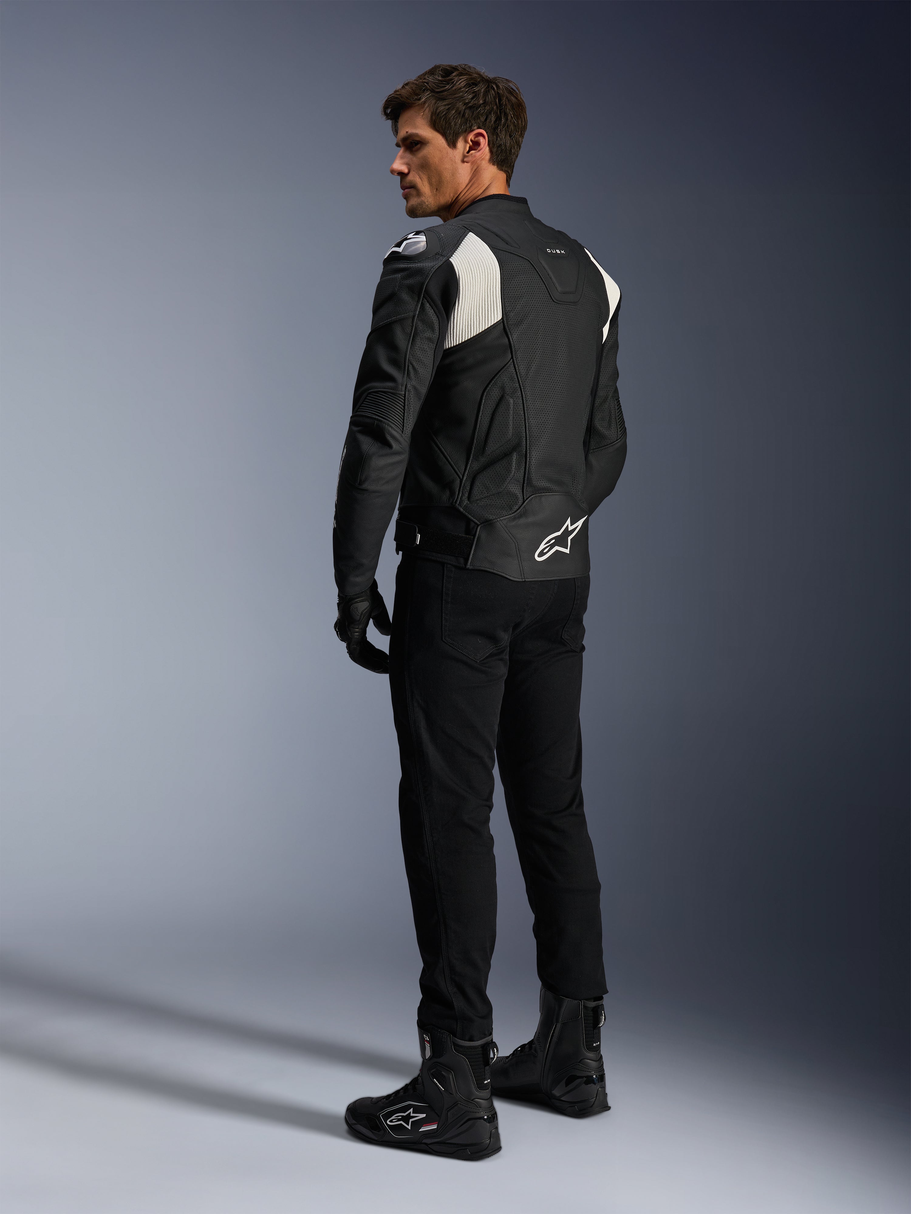 Giacca in pelle Alpinestars Dusk Airflow, nero e bianco, vista posteriore del modello che indossa la giacca in pelle traforata con pantaloni e stivali neri coordinati, caratterizzata da pannelli elastici a fisarmonica bianchi e logo sulla parte bassa della schiena, abbigliamento tecnico da moto