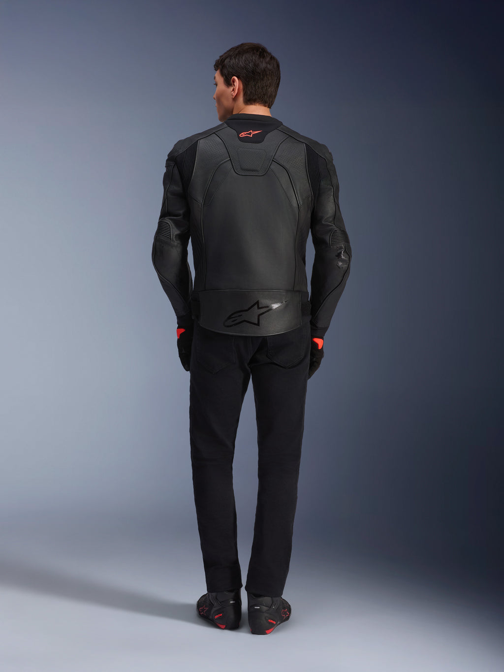 Alpinestars GP Plus R V4 Airflow Leather Jacket, nero, vista posteriore del pilota con equipaggiamento completo, pelle traforata per la ventilazione e accenti con logo rosso, design sportivo aderente, abbinata a pantaloni neri e stivali da moto
