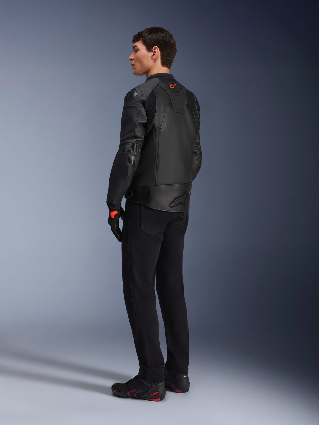 Giacca in pelle Alpinestars GP Plus R V4 Airflow, giacca da moto sportiva da uomo, completamente nera, vista posteriore a tre quarti su un modello, con pannelli in pelle traforata, logo rosso sul retro e protezioni integrate sulle spalle, indossata con pantaloni neri e scarpe da guida