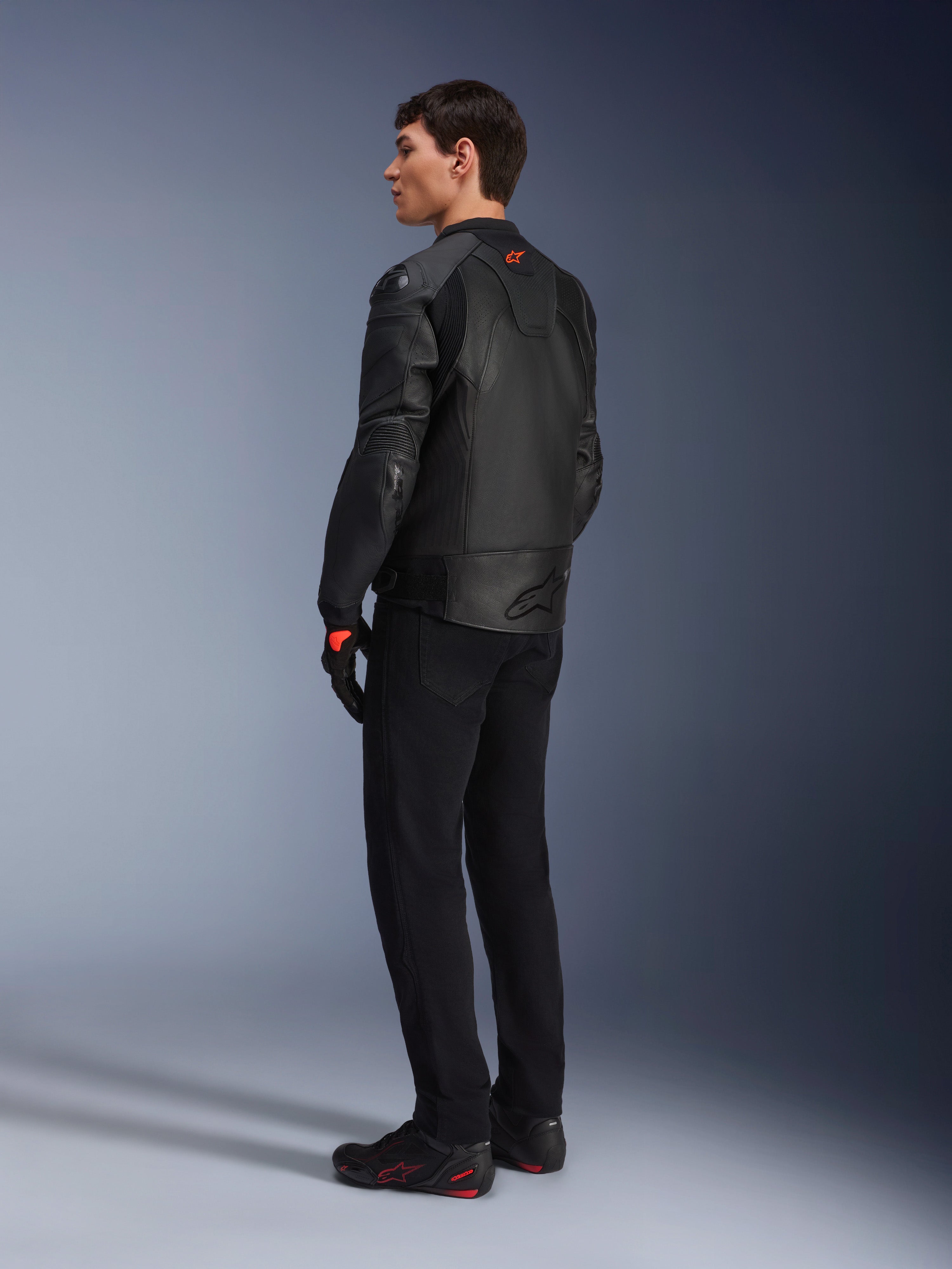 Giacca in pelle Alpinestars GP Plus R V4 Airflow, giacca da moto sportiva da uomo, completamente nera, vista posteriore a tre quarti su un modello, con pannelli in pelle traforata, logo rosso sul retro e protezioni integrate sulle spalle, indossata con pantaloni neri e scarpe da guida
