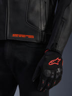 Giacca in pelle Alpinestars GP Plus R V4 Airflow, primo piano della pelle nera traforata con scritte grafiche arancioni, abbinata a un guanto da moto nero e arancione con logo Astars