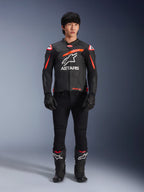 Modello che indossa la giacca in pelle Alpinestars GP Plus R V4 Airflow, nera con accenti rosso fluo e bianchi, grande logo Astars bianco sul petto, abbinata a pantaloni tecnici, guanti e stivali neri, abbigliamento da moto ad alte prestazioni in studio