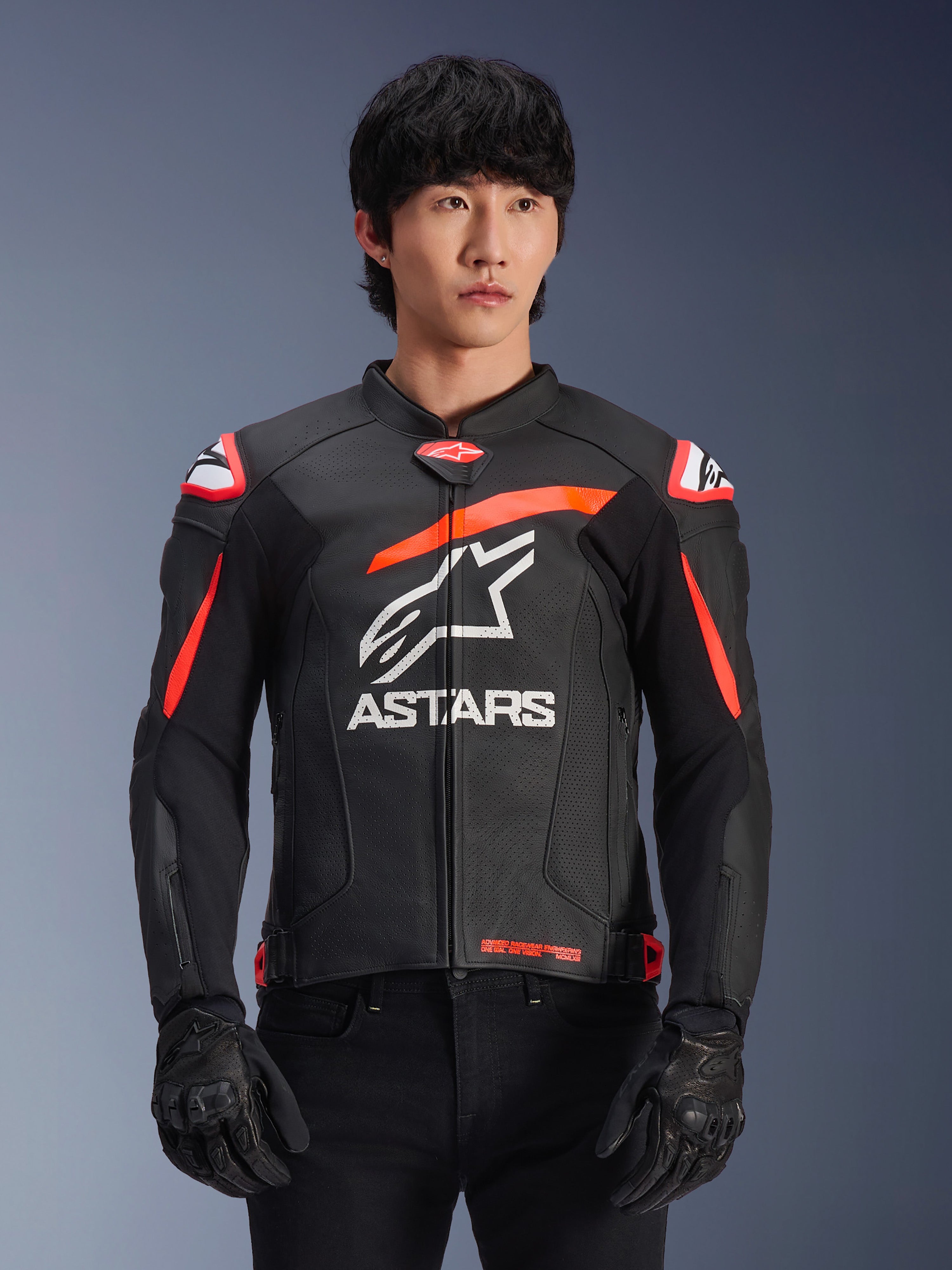 Alpinestars GP Plus R V4 Airflow Leather Jacket in nero con accenti rosso fluo e bianchi, indossata da un modello, con costruzione in pelle traforata, un grande logo ASTARS bianco sul petto e protezioni per le spalle integrate per il motociclismo racing.
