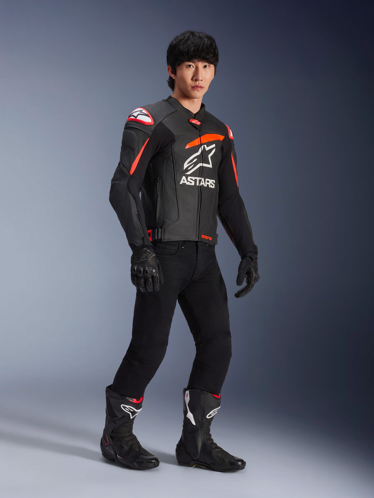 Pilota che indossa la giacca in pelle Alpinestars GP Plus R V4 Airflow, nero con accenti rosso fluo e bianco, con pelle traforata e logo ASTARS sul petto, abbinata a pantaloni da moto neri e stivali da moto neri, abbigliamento sport performance
