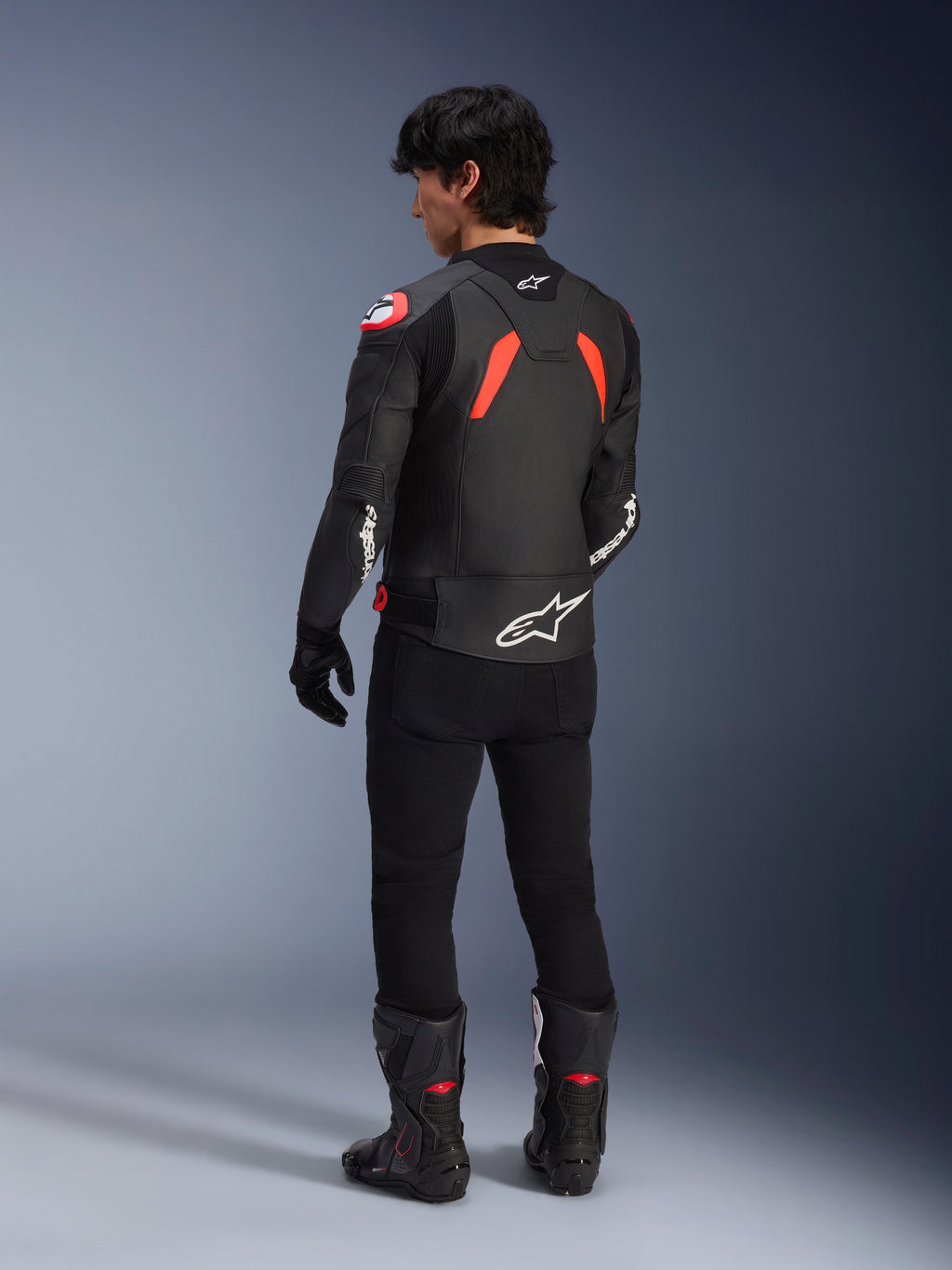 Vista posteriore di un modello che indossa la giacca in pelle Alpinestars GP Plus R V4 Airflow, colore nero con accenti rosso fluo e bianchi, abbinata a pantaloni neri e stivali da moto neri, con loghi bianchi evidenti sulla schiena e sulle maniche, costruzione in pelle in stile sportivo