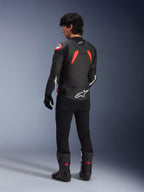 Vista posteriore di un modello che indossa la giacca in pelle Alpinestars GP Plus R V4 Airflow, colore nero con accenti rosso fluo e bianchi, abbinata a pantaloni neri e stivali da moto neri, con loghi bianchi evidenti sulla schiena e sulle maniche, costruzione in pelle in stile sportivo