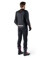 Giacca in pelle Alpinestars Faster V3 Airflow, nero e rosso fluo, vista da dietro su un pilota, con pannelli in pelle traforata e loghi evidenti, indossata con pantaloni neri e stivali da moto, abbigliamento da guida sportiva.