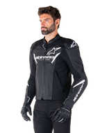 Giacca in pelle Alpinestars Faster V3 Airflow, nero e bianco, giacca da moto sportiva con pannelli in pelle traforata, loghi Alpinestars bianchi su petto e braccia, indossata da un modello con guanti e pantaloni neri, abbigliamento tecnico protettivo