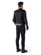 Vista posteriore di un pilota che indossa la giacca in pelle Alpinestars Faster V3 Airflow, nero con dettagli bianchi e giallo fluo, abbinata a pantaloni e stivali neri, con pelle traforata per la ventilazione e il marchio 'RACING AHEAD' sulla parte bassa della schiena.