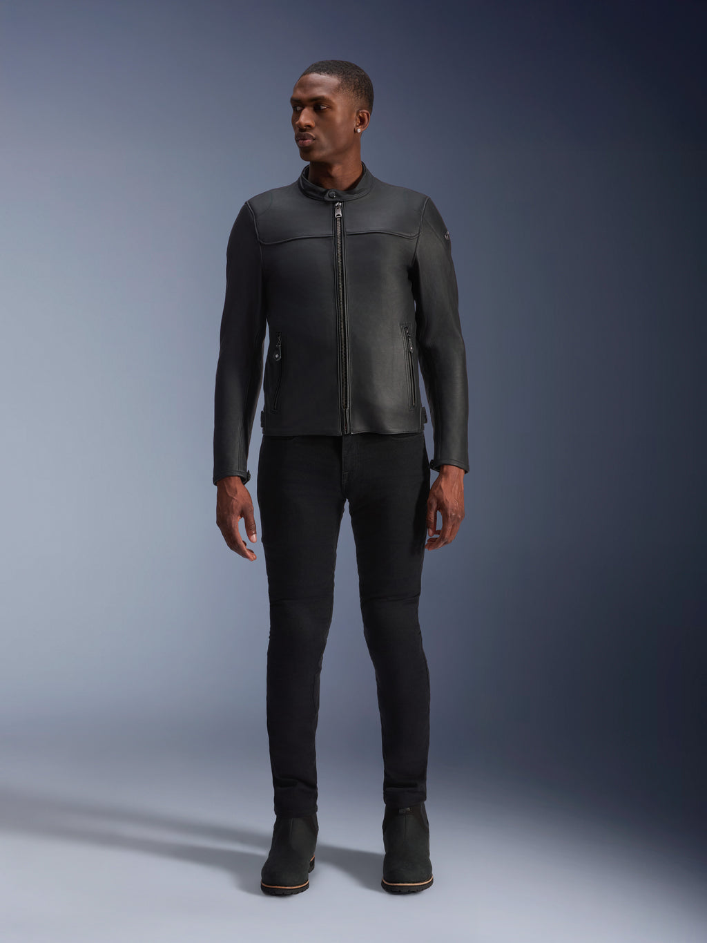 Modello che indossa la giacca in pelle Alpinestars Blacktrack, pelle bovina premium nera, design da motociclista moderno e minimalista con vestibilità su misura, abbinata a pantaloni da moto e stivali neri, stile urbano contemporaneo