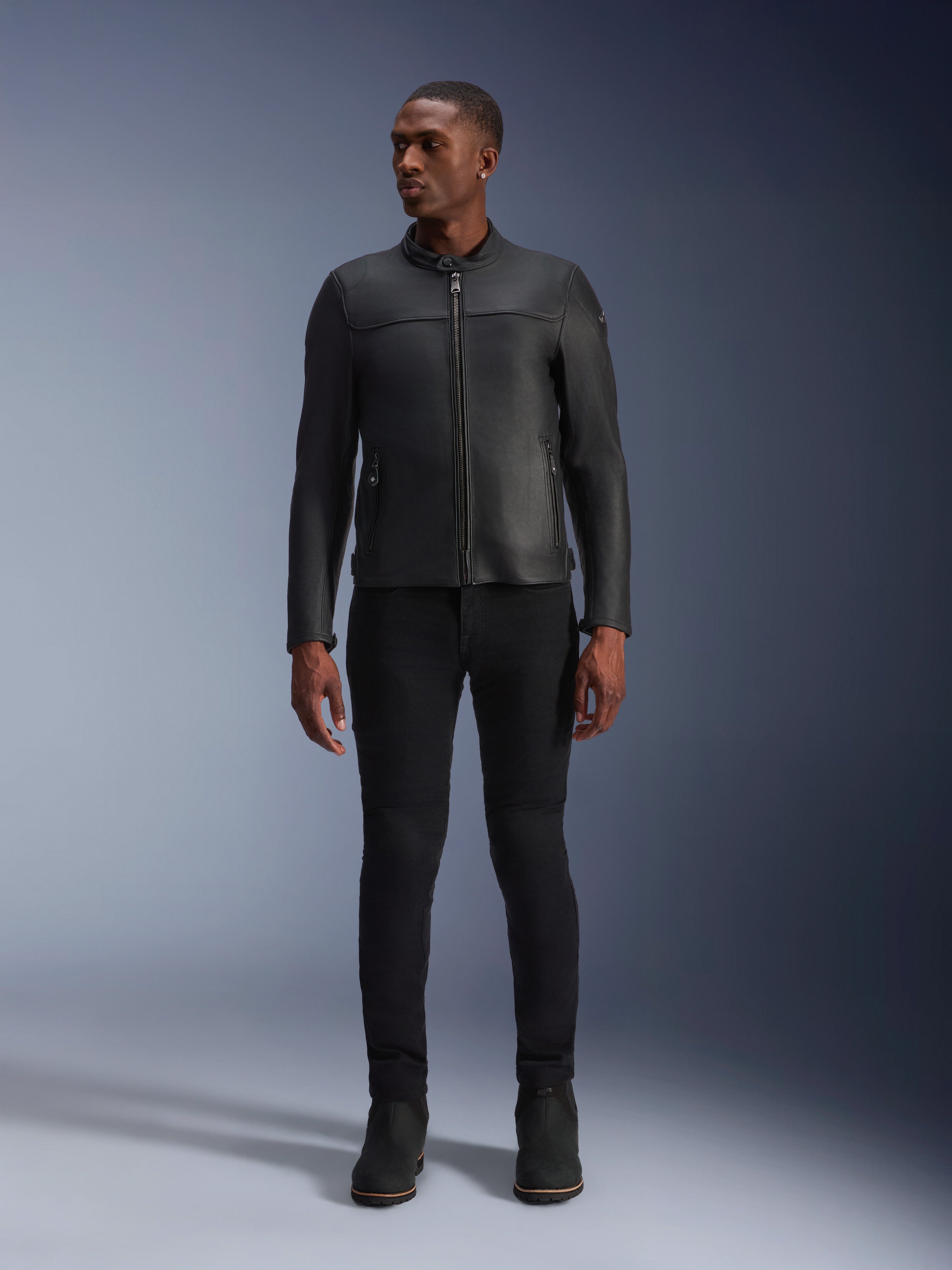 Modello che indossa la giacca in pelle Alpinestars Blacktrack, pelle bovina premium nera, design da motociclista moderno e minimalista con vestibilità su misura, abbinata a pantaloni da moto e stivali neri, stile urbano contemporaneo