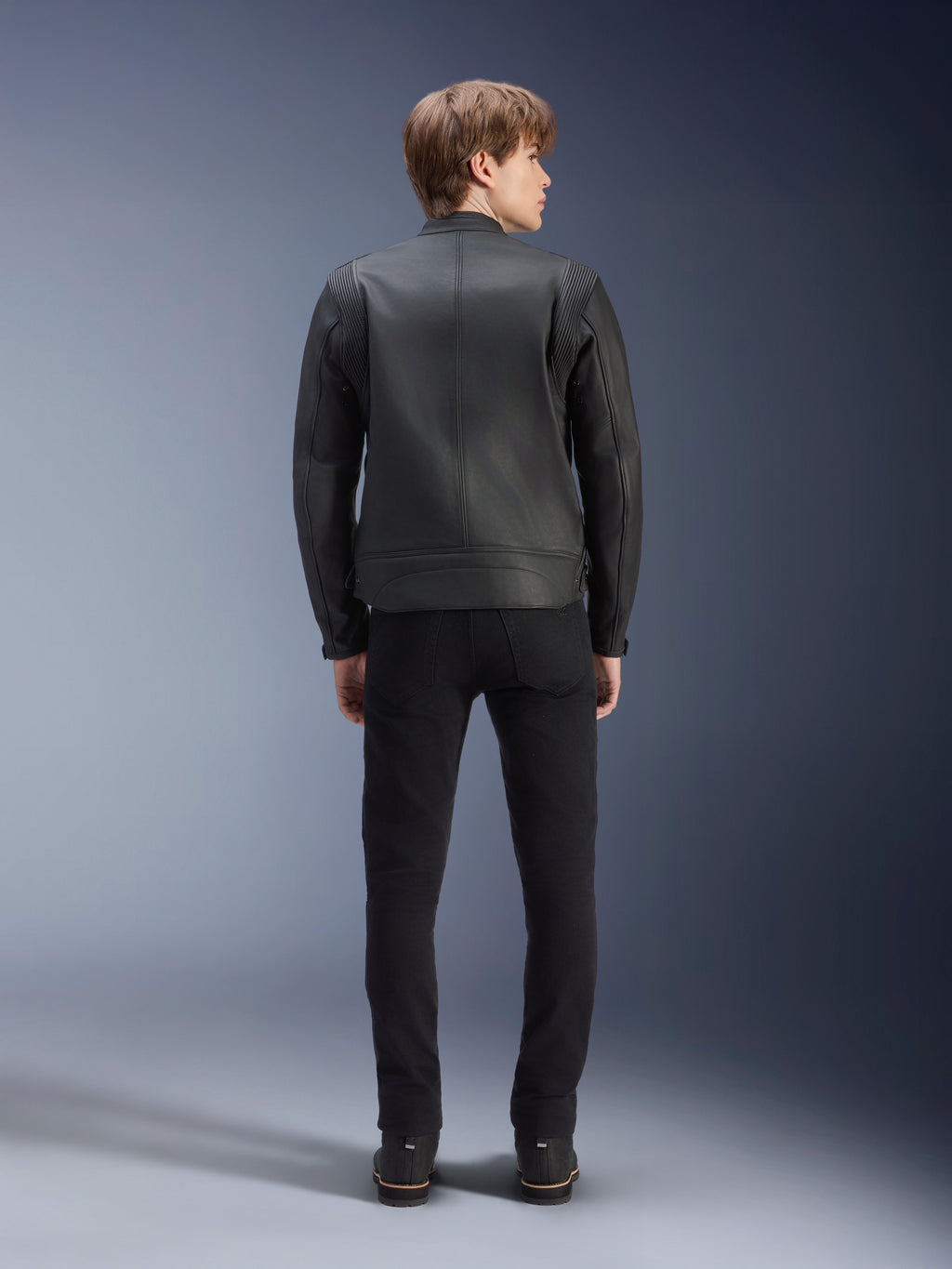 Giacca in pelle Alpinestars Dyno, nera, vista posteriore su modello, pelle bovina premium con pannelli a fisarmonica sulle spalle, giacca da moto d'ispirazione heritage, indossata con pantaloni e stivali neri, stile urban contemporaneo
