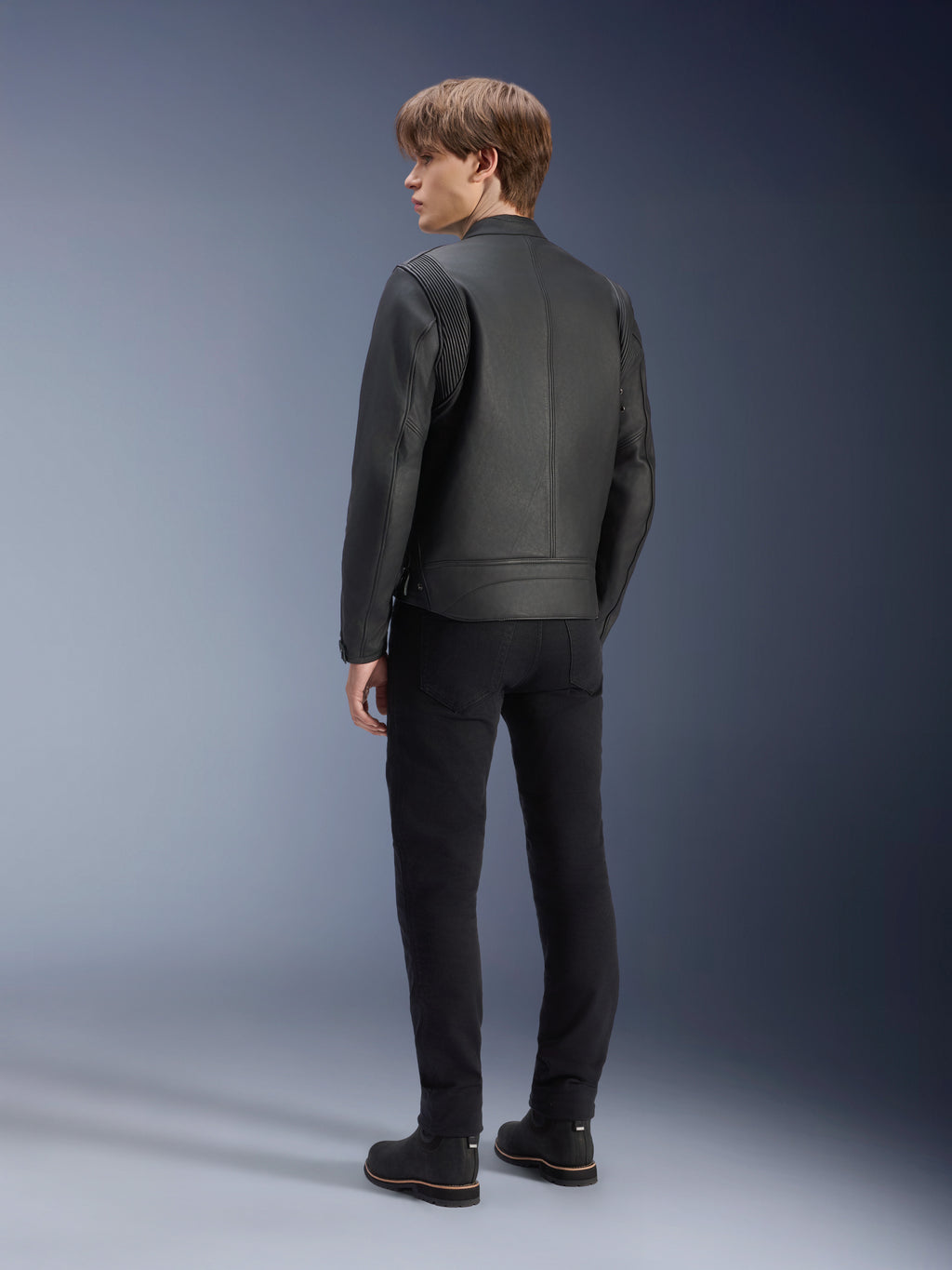 Giacca in pelle Alpinestars Dyno, nero, indossata da un modello uomo, vista posteriore, con pannelli a coste sulle spalle, abbinata a pantaloni e stivali neri, stile motociclistico urbano d'ispirazione heritage