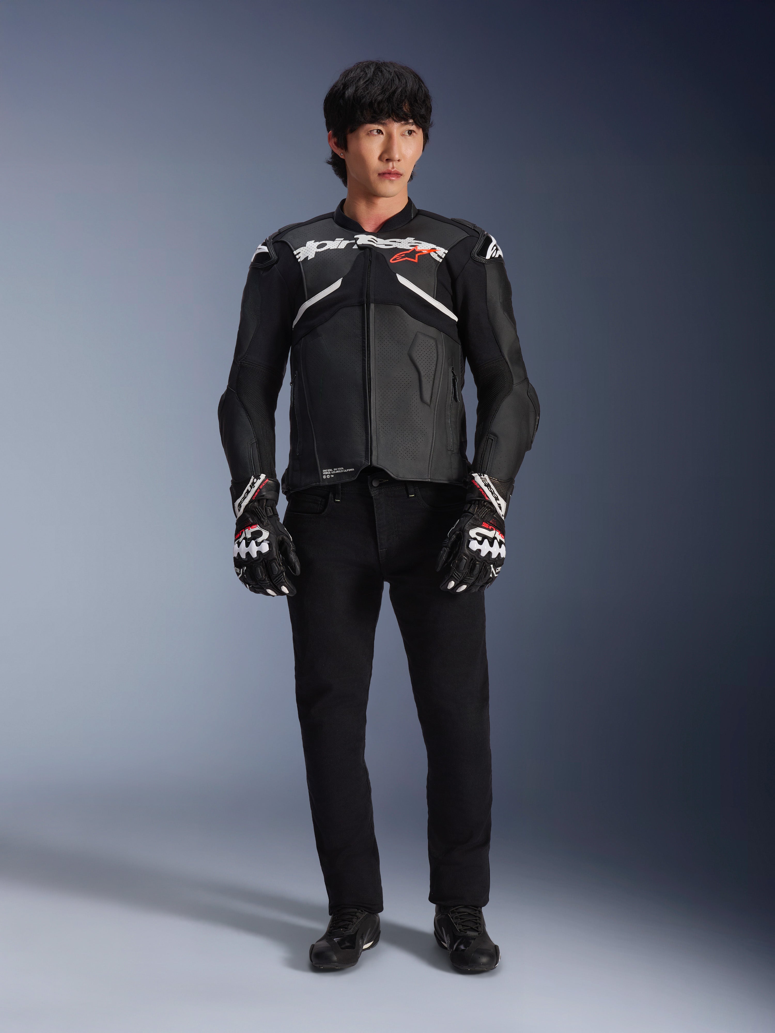 Modello che indossa la giacca in pelle Alpinestars Atem V5 in nero e bianco, con marchio Alpinestars bianco e logo con stella rossa sul petto, abbinata a pantaloni neri e guanti da corsa bianchi e neri, abbigliamento da moto performante ed elegante