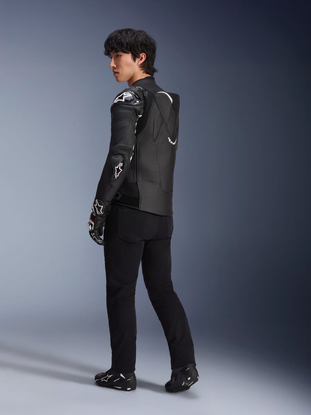 Modello che indossa la giacca in pelle Alpinestars Atem V5 in nero e bianco, vista posteriore a tre quarti che mostra la gobba aerodinamica e le protezioni DFS RR, abbinata a pantaloni neri, guanti racing e stivali, abbigliamento professionale da moto