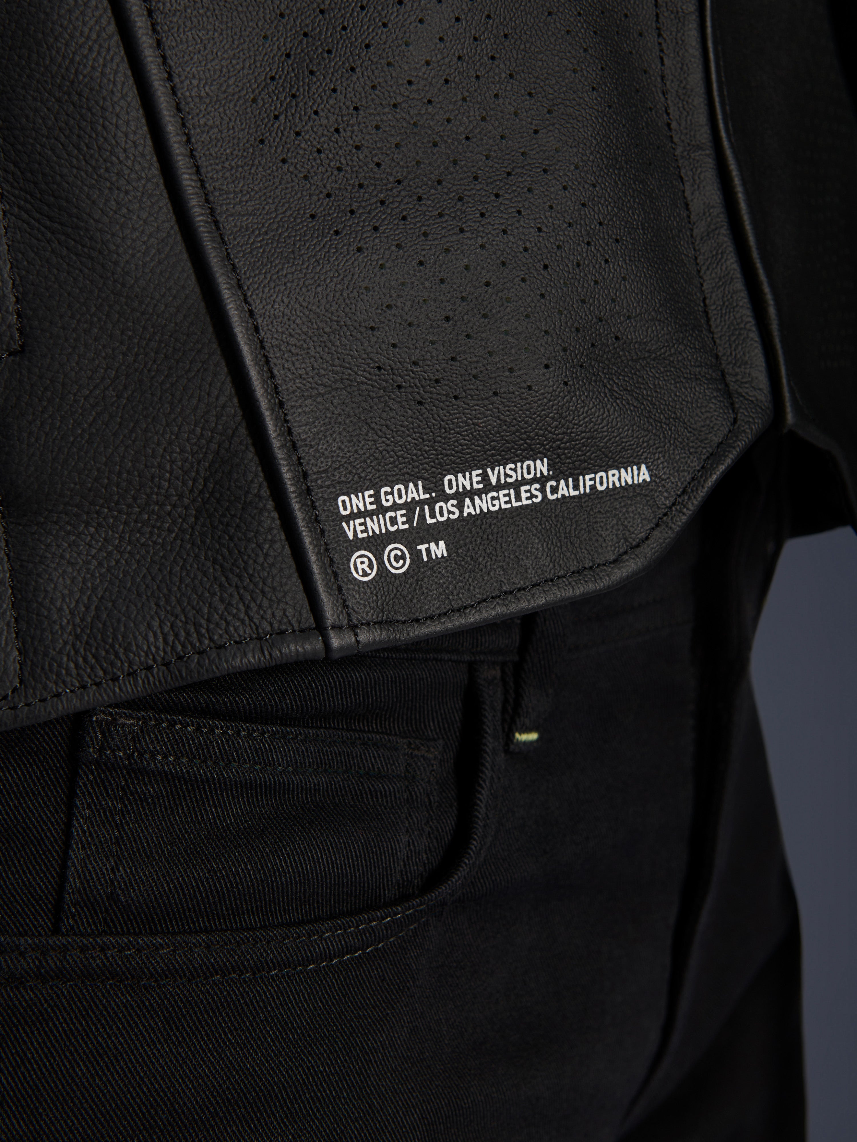 Primo piano della giacca in pelle Alpinestars Atem V5 in nero, con pannello posteriore inferiore, marchio 'ONE GOAL. ONE VISION. VENICE / LOS ANGELES CALIFORNIA' e trama in pelle traforata.