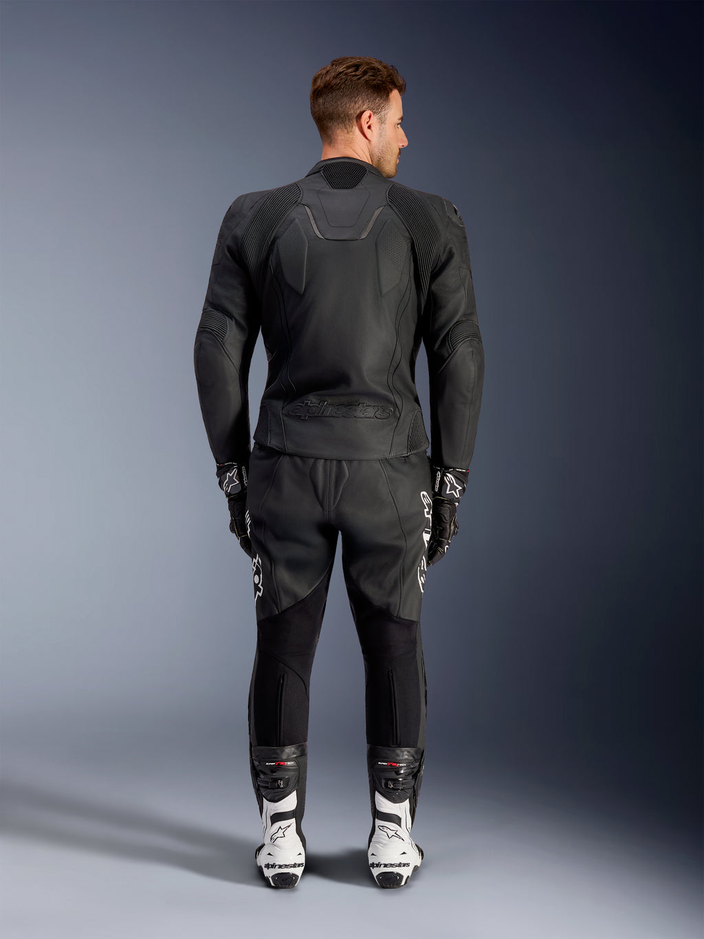 Vista posteriore di un uomo che indossa la giacca in pelle Alpinestars Caliber in nero, con logo in rilievo e pannelli in pelle traforata, abbinata a pantaloni da moto in pelle neri, guanti neri e stivali racing bianchi.
