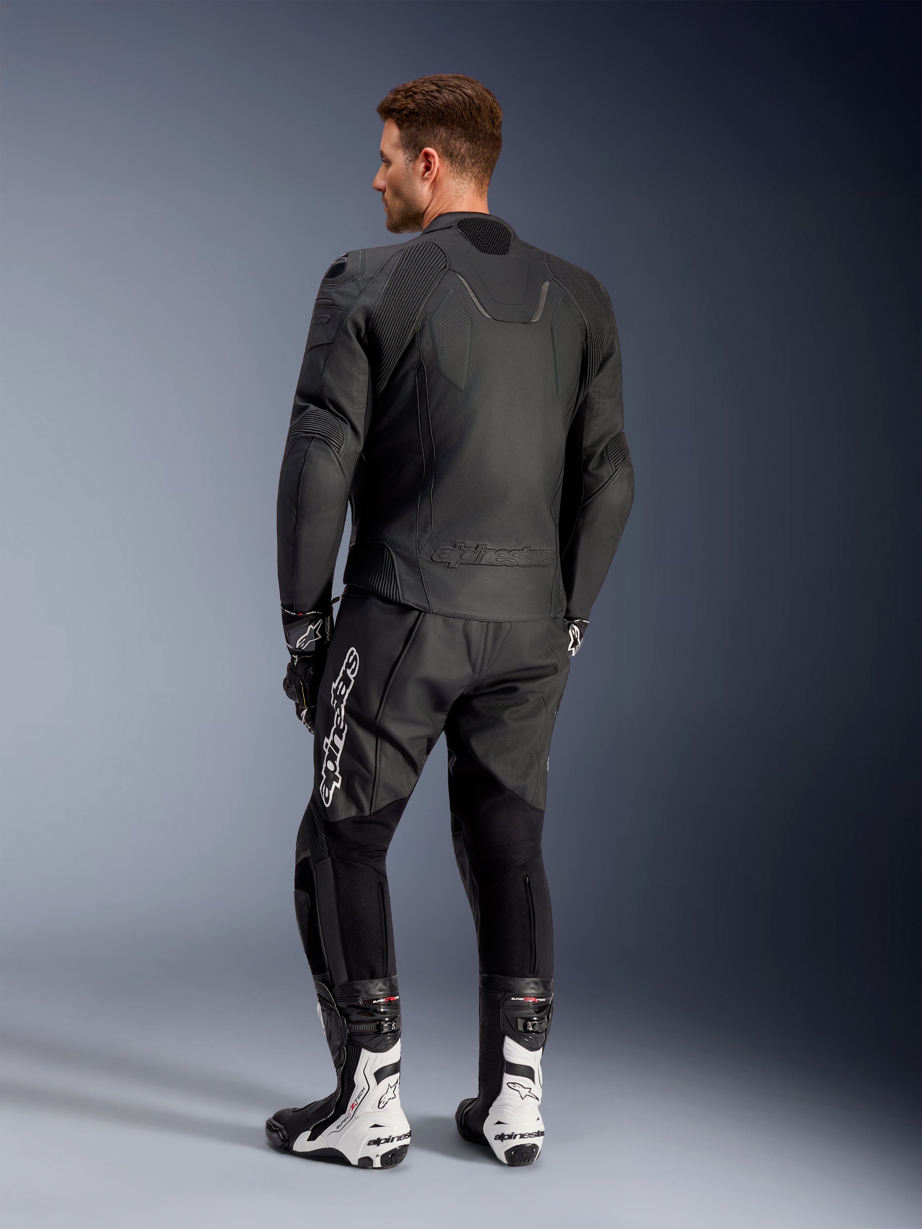 Giacca in pelle Alpinestars Caliber, giacca da moto stradale nera, vista posteriore, con pelle traforata e inserti a soffietto elasticizzati su vita e spalle, logo a rilievo, indossata con pantaloni in pelle neri e stivali da corsa bianchi