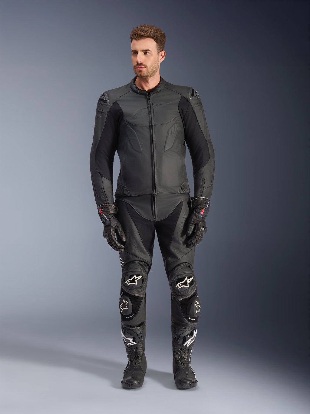 Giacca in pelle Alpinestars Caliber, giacca da moto nera, indossata con pantaloni da corsa, guanti e stivali coordinati, caratterizzata da un design elegante con loghi tono su tono, pannelli elasticizzati sulle maniche e slider protettivi sulle spalle, ambientazione in studio professionale