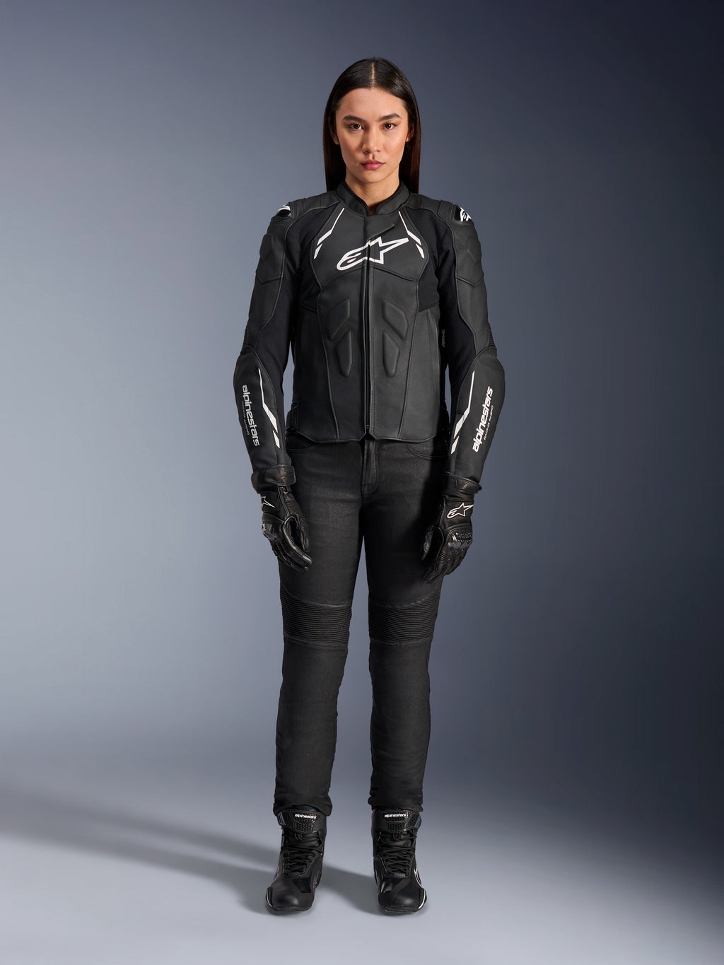Giacca in pelle Alpinestars Stella Dusk da donna, nero e bianco, indossata da una modella con pantaloni e stivali neri coordinati, vista frontale, con loghi Alpinestars bianchi sul petto e sulle maniche, equipaggiamento protettivo in pregiata pelle bovina per donna