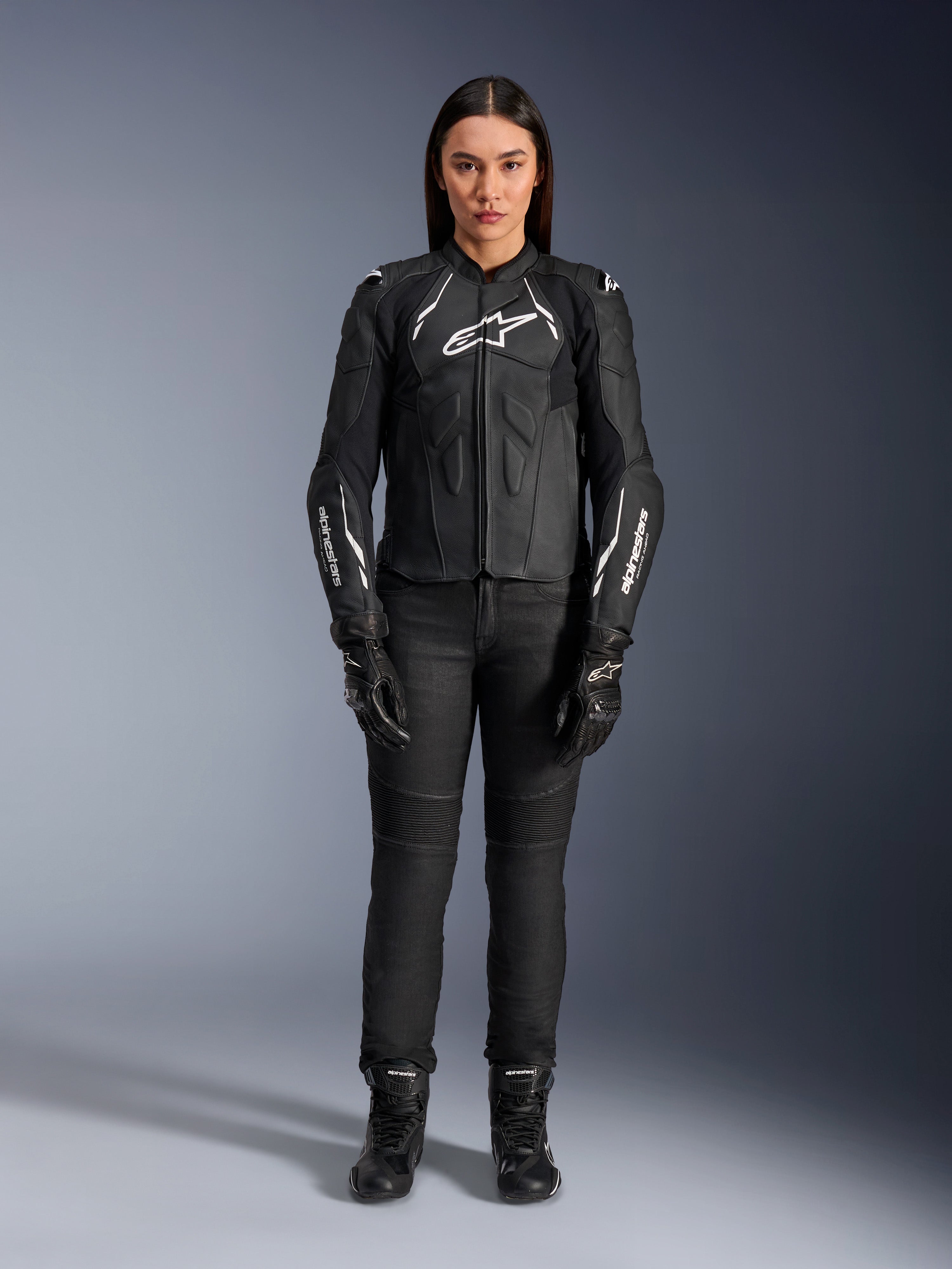 Giacca in pelle Alpinestars Stella Dusk da donna, nero e bianco, indossata da una modella con pantaloni e stivali neri coordinati, vista frontale, con loghi Alpinestars bianchi sul petto e sulle maniche, equipaggiamento protettivo in pregiata pelle bovina per donna