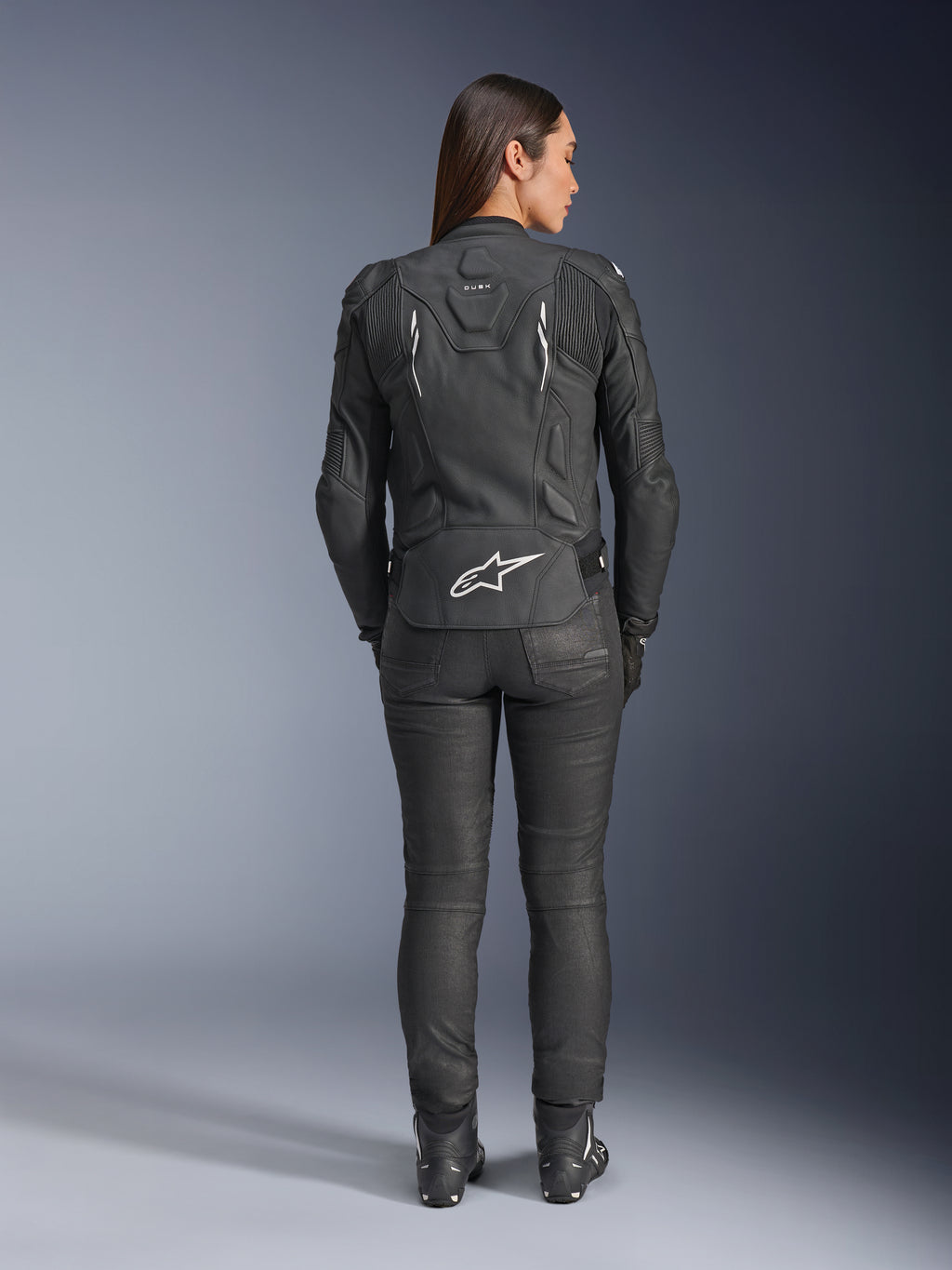 Giacca in pelle Alpinestars Stella Dusk da donna, nero, vista posteriore con logo Alpinestars bianco sulla parte bassa della schiena e marchio 'DUSK' sulla parte superiore, indossata con pantaloni e stivali da moto neri, mette in risalto la vestibilità femminile sagomata e la struttura protettiva in pelle