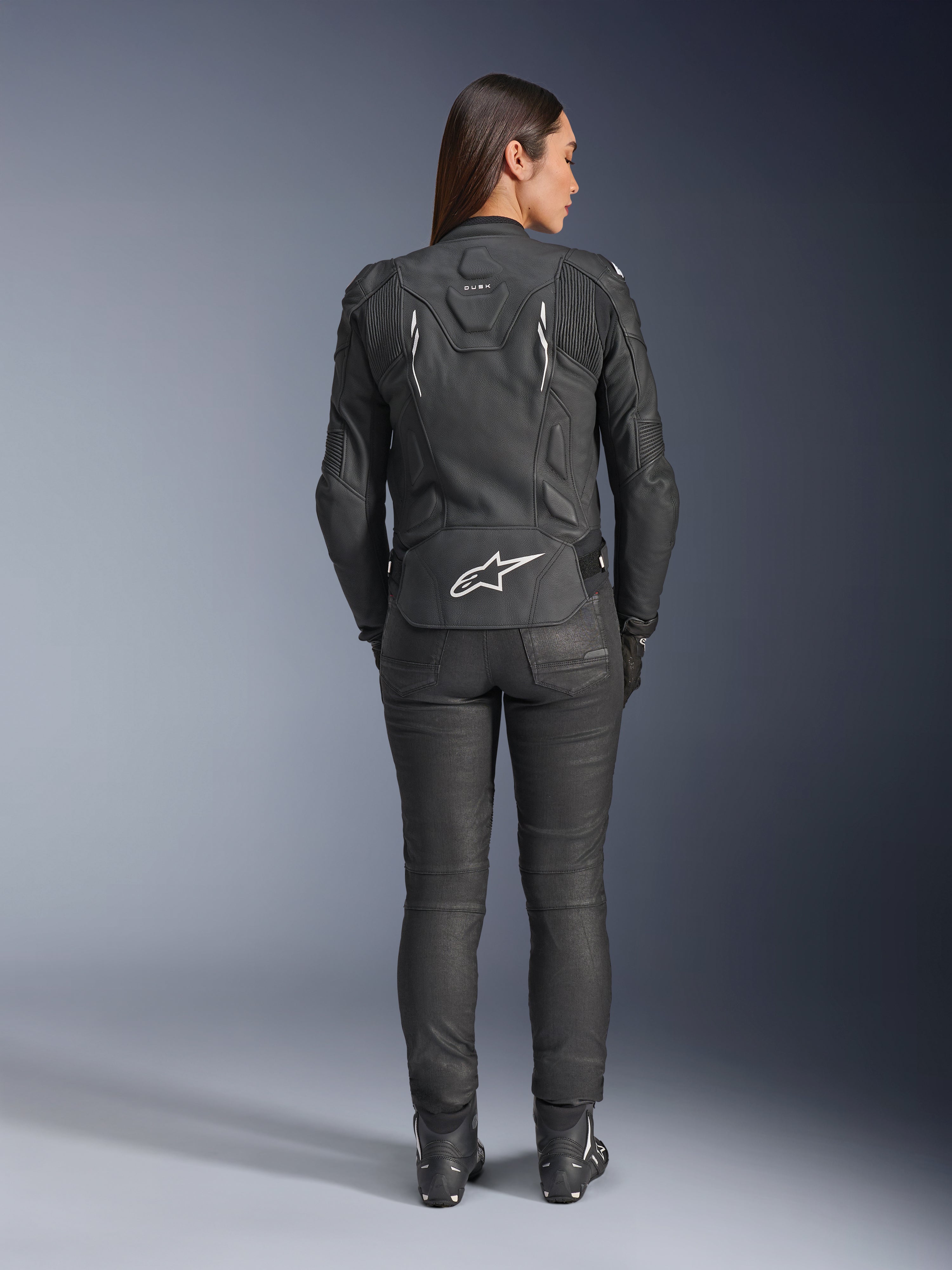 Giacca in pelle Alpinestars Stella Dusk da donna, nero, vista posteriore con logo Alpinestars bianco sulla parte bassa della schiena e marchio 'DUSK' sulla parte superiore, indossata con pantaloni e stivali da moto neri, mette in risalto la vestibilità femminile sagomata e la struttura protettiva in pelle