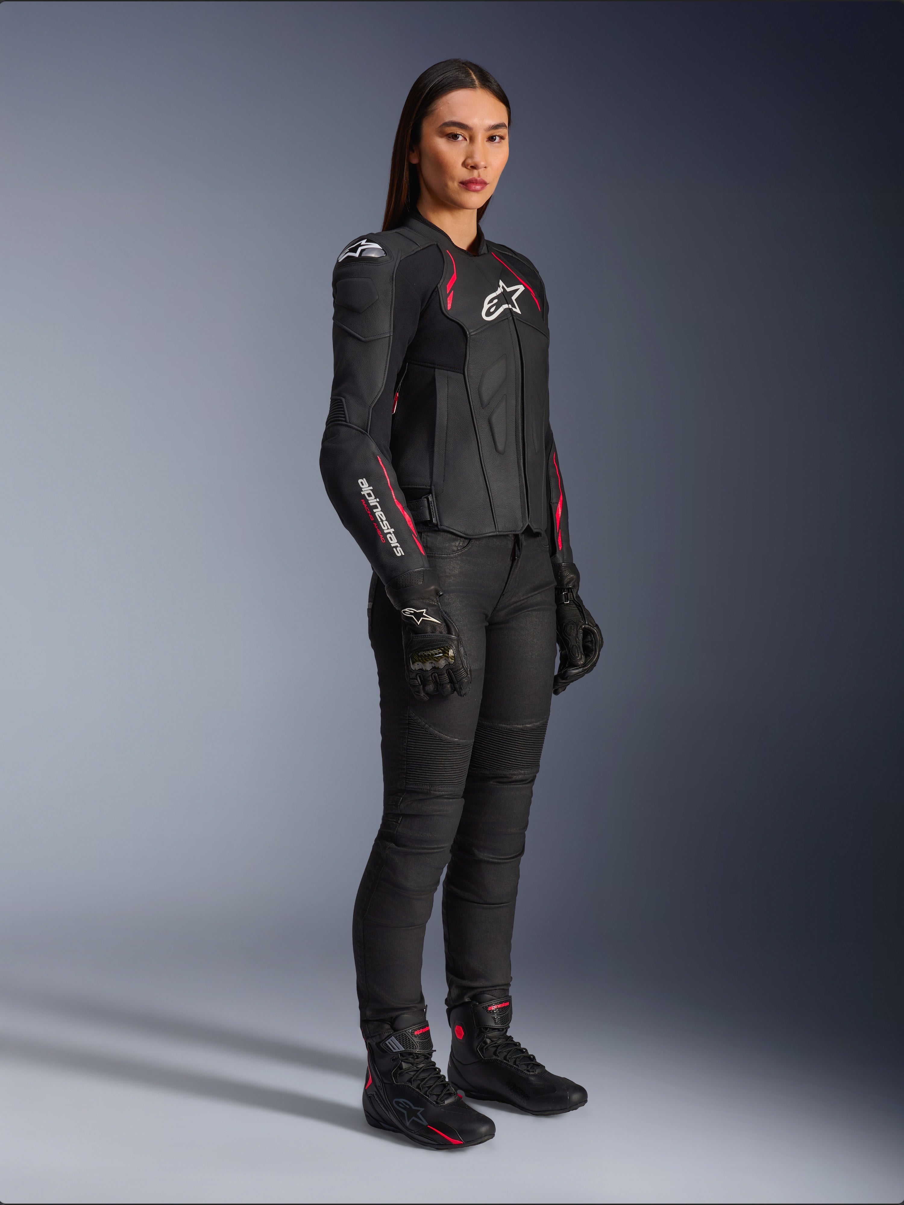 Alpinestars Stella Dusk Leather Jacket da donna, nero, rosa Diva e bianco, indossata da una modella con pantaloni e stivali da moto neri, struttura in pelle con dettagli rosa e loghi bianchi