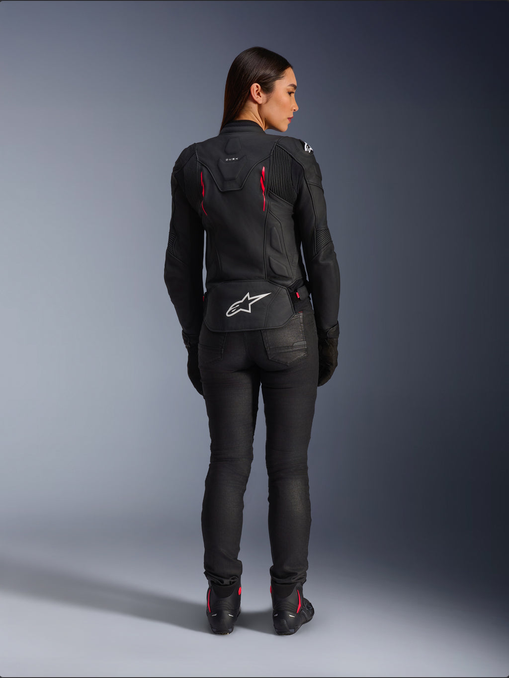 Giacca in pelle Alpinestars Stella Dusk, vista posteriore, pelle nera con accenti Diva Pink e bianchi, presenta il logo Alpinestars bianco sulla parte bassa della schiena e dettagli delle prese d'aria rosa, indossata con pantaloni da moto e stivali neri, abbigliamento sportivo da moto premium per donna