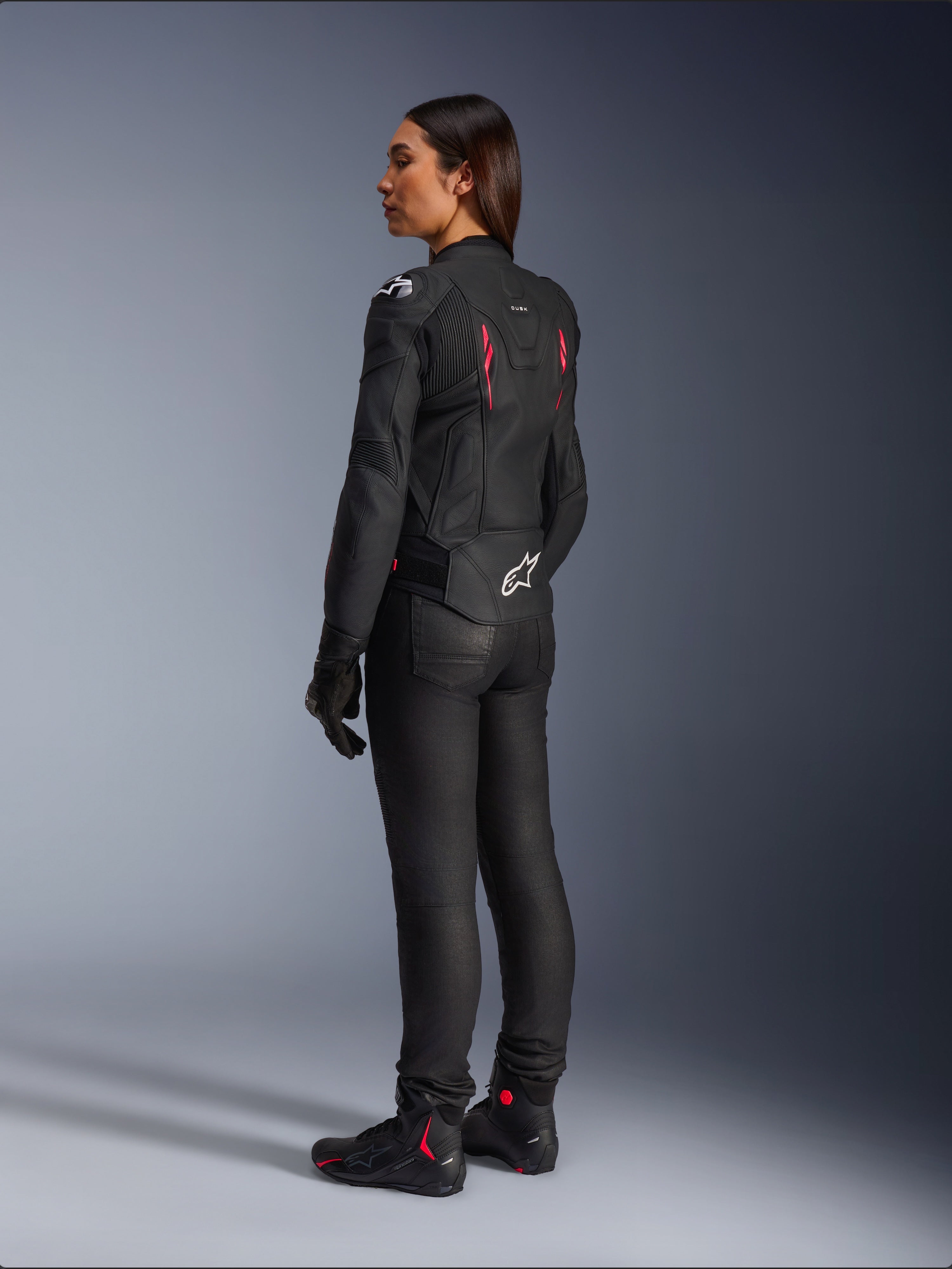Giacca in pelle da donna Alpinestars Stella Dusk, Nero Diva Pink Bianco, vista posteriore di tre quarti su una pilota, indossata con pantaloni da moto neri e stivali neri, elegante struttura in pelle con accenti rosa e logo Alpinestars bianco sulla parte bassa della schiena
