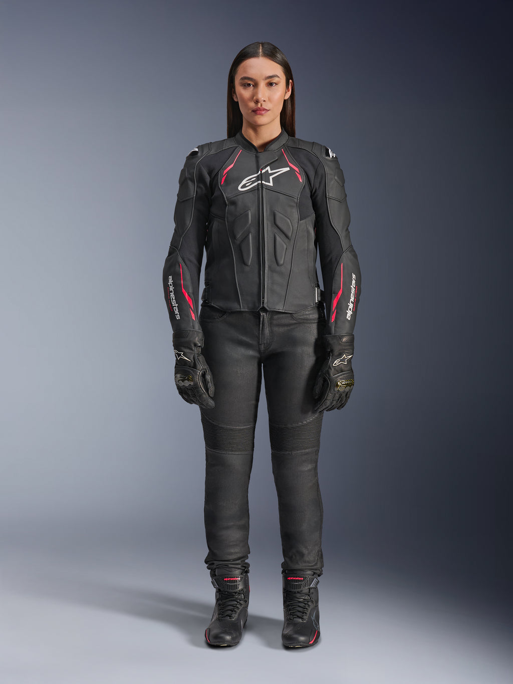 Una donna indossa la giacca in pelle Alpinestars Stella Dusk in Black Diva Pink White, con logo bianco sul petto e accenti rosa su spalle e maniche, abbinata a pantaloni da moto, guanti e stivali neri.