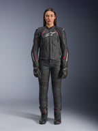 Una donna indossa la giacca in pelle Alpinestars Stella Dusk in Black Diva Pink White, con logo bianco sul petto e accenti rosa su spalle e maniche, abbinata a pantaloni da moto, guanti e stivali neri.
