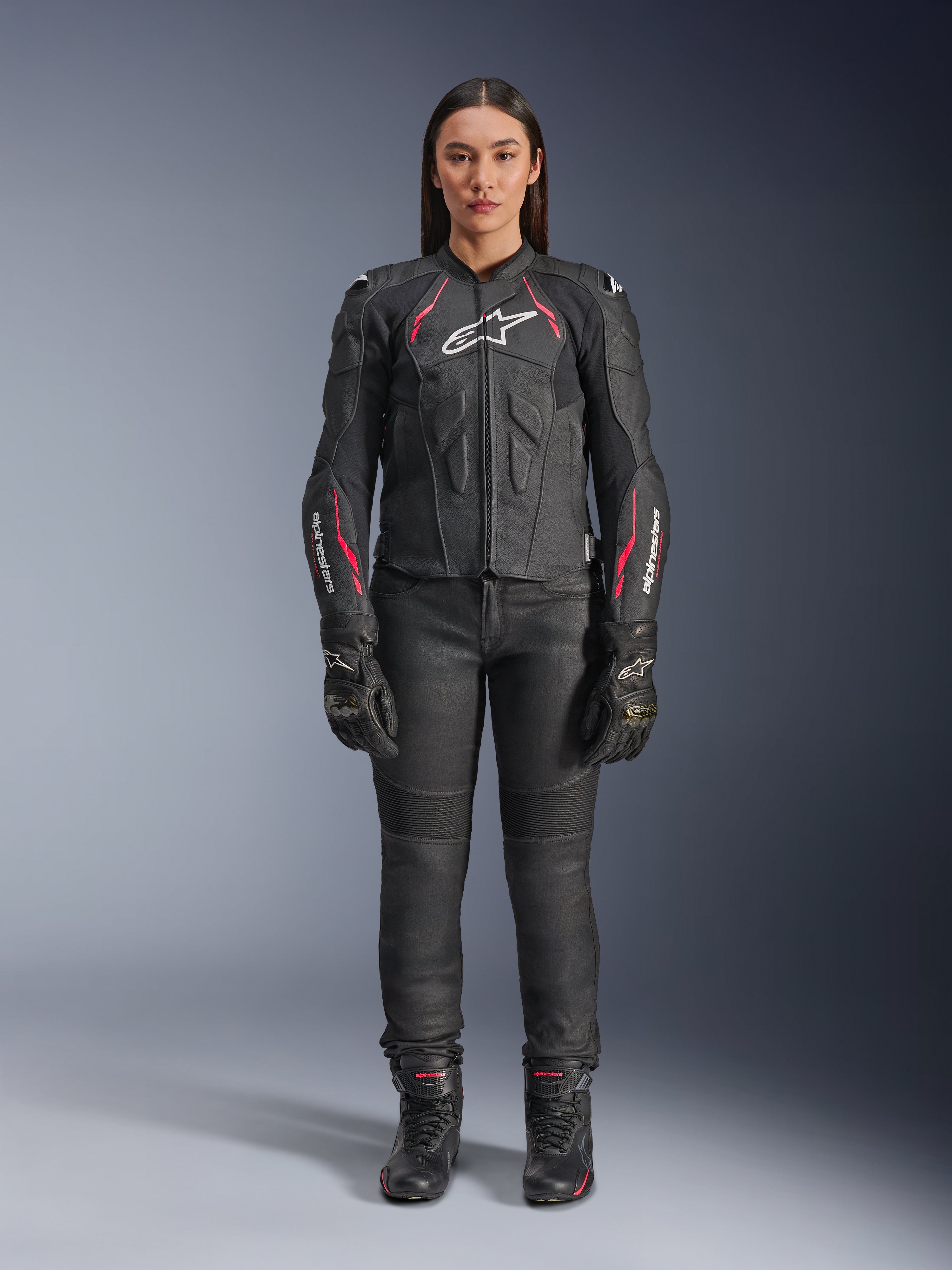 Una donna indossa la giacca in pelle Alpinestars Stella Dusk in Black Diva Pink White, con logo bianco sul petto e accenti rosa su spalle e maniche, abbinata a pantaloni da moto, guanti e stivali neri.
