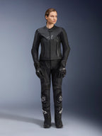 Giacca in pelle Alpinestars Stella GP Plus V4 da donna in nero, indossata da una motociclista con pantaloni racing in pelle, guanti e stivali coordinati, caratterizzata da una vestibilità sagomata, cerniera centrale e discreto logo sul petto, coordinata per la guida sportiva e su strada.