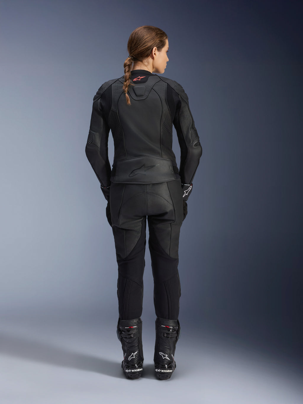 Alpinestars Giacca in pelle Stella GP Plus V4 da donna, Nero Nero, vista posteriore a figura intera di una motociclista con pantaloni in pelle e stivali coordinati, design minimalista con un piccolo logo rosso sul collo, silhouette sportiva sagomata per donna