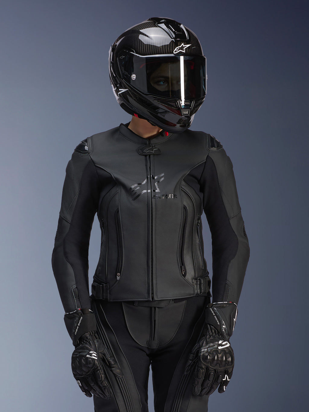 Giacca in pelle Alpinestars Stella GP Plus V4 da donna in nero, indossata con casco in fibra di carbonio e guanti racing, costruzione in pelle elegante con logo discreto sul petto, silhouette sagomata per motocicliste, abbigliamento moto sportivo professionale