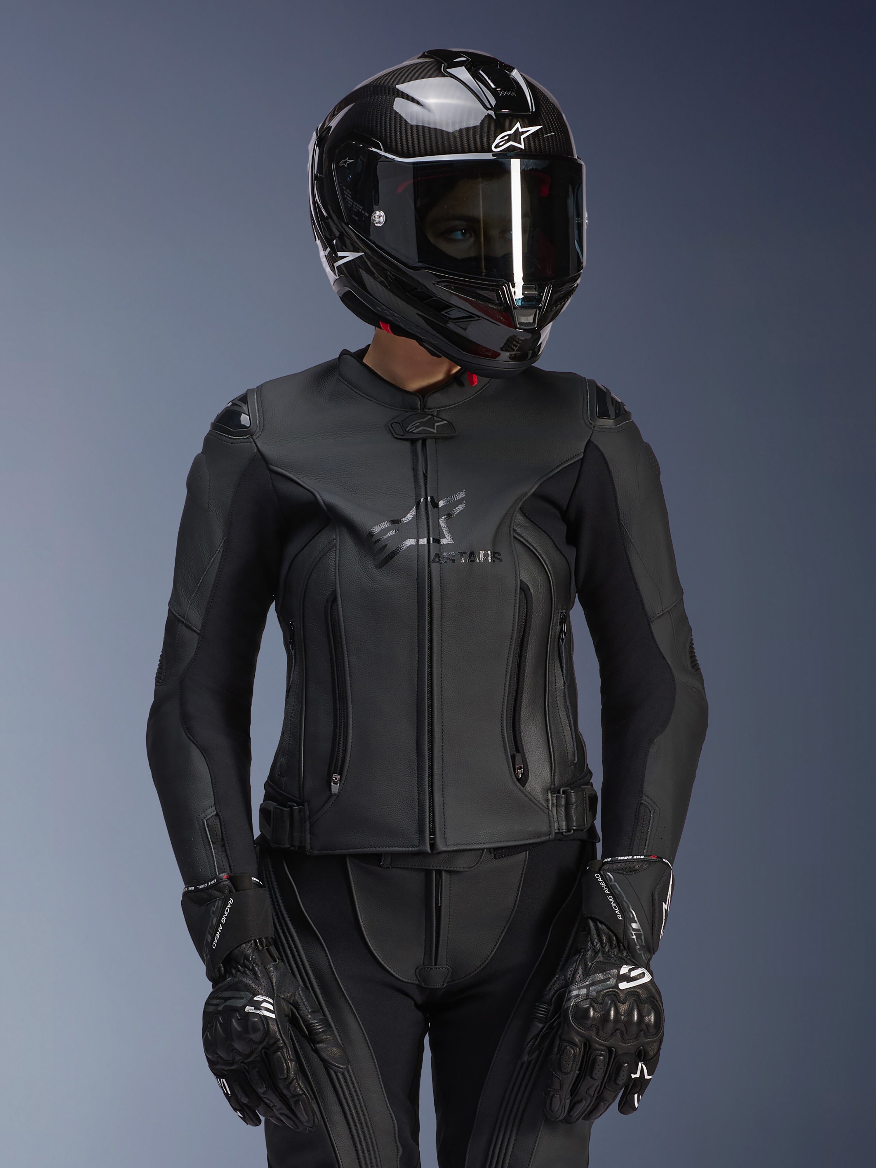 Giacca in pelle Alpinestars Stella GP Plus V4 da donna in nero, indossata con casco in fibra di carbonio e guanti racing, costruzione in pelle elegante con logo discreto sul petto, silhouette sagomata per motocicliste, abbigliamento moto sportivo professionale