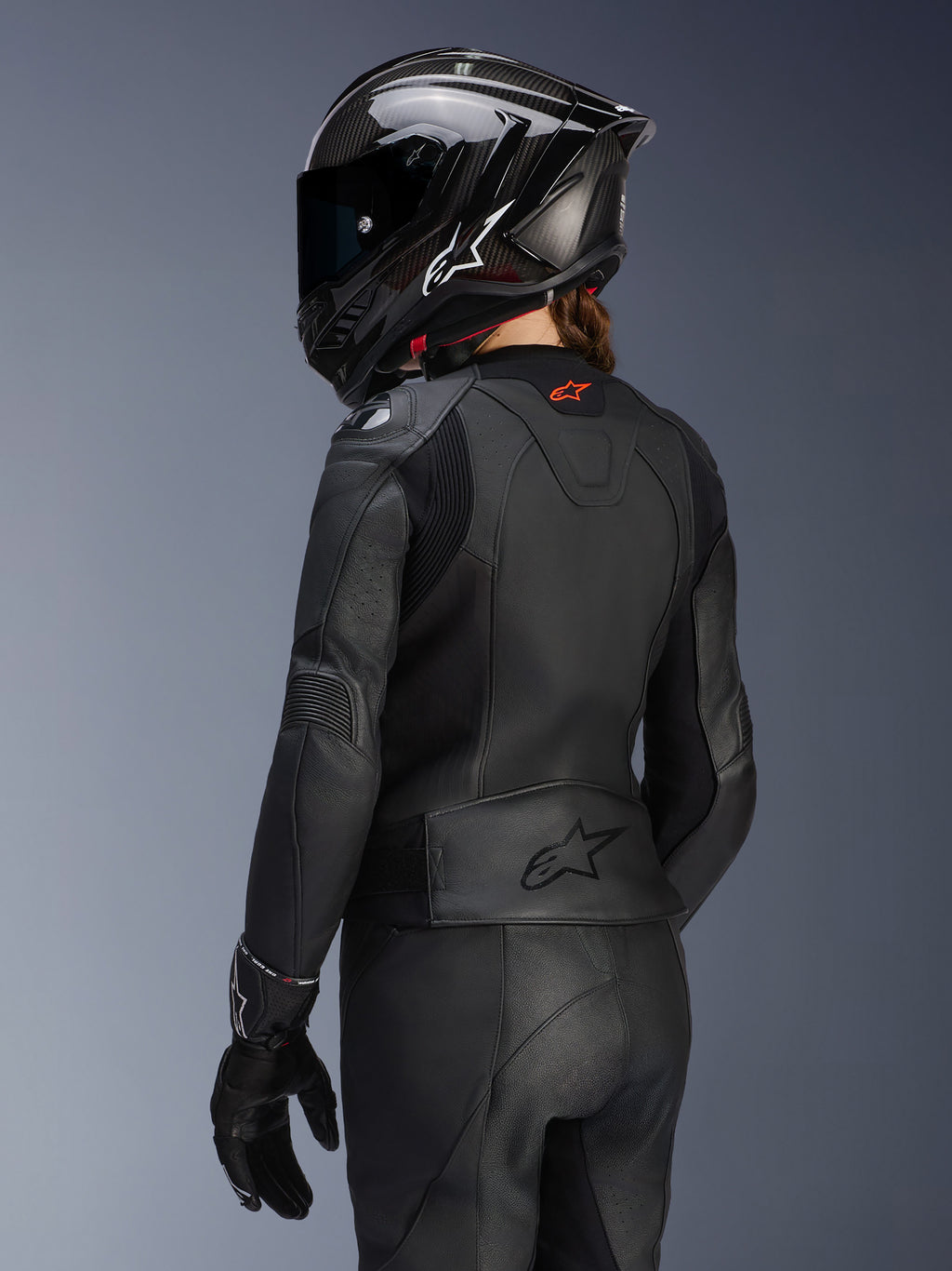 Motociclista vista da dietro che indossa la giacca in pelle Alpinestars Stella GP Plus V4 da donna in nero, pantaloni e guanti in pelle coordinati e un casco integrale nero stile carbonio, mettendo in risalto la vestibilità aerodinamica della giacca e il logo goffrato sulla parte bassa della schiena.