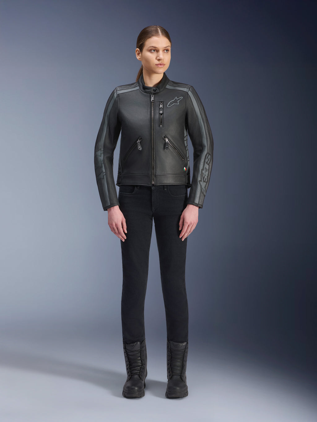 Alpinestars Stella Dyno Leather Jacket, giacca da moto da donna, nero, grigio catrame e grigio scuro, vista in studio a figura intera di una modella che indossa la giacca con pantaloni neri slim-fit e stivali neri, realizzata in pelle bovina con accenti grigi e logo Alpinestars sul petto e sulle maniche.