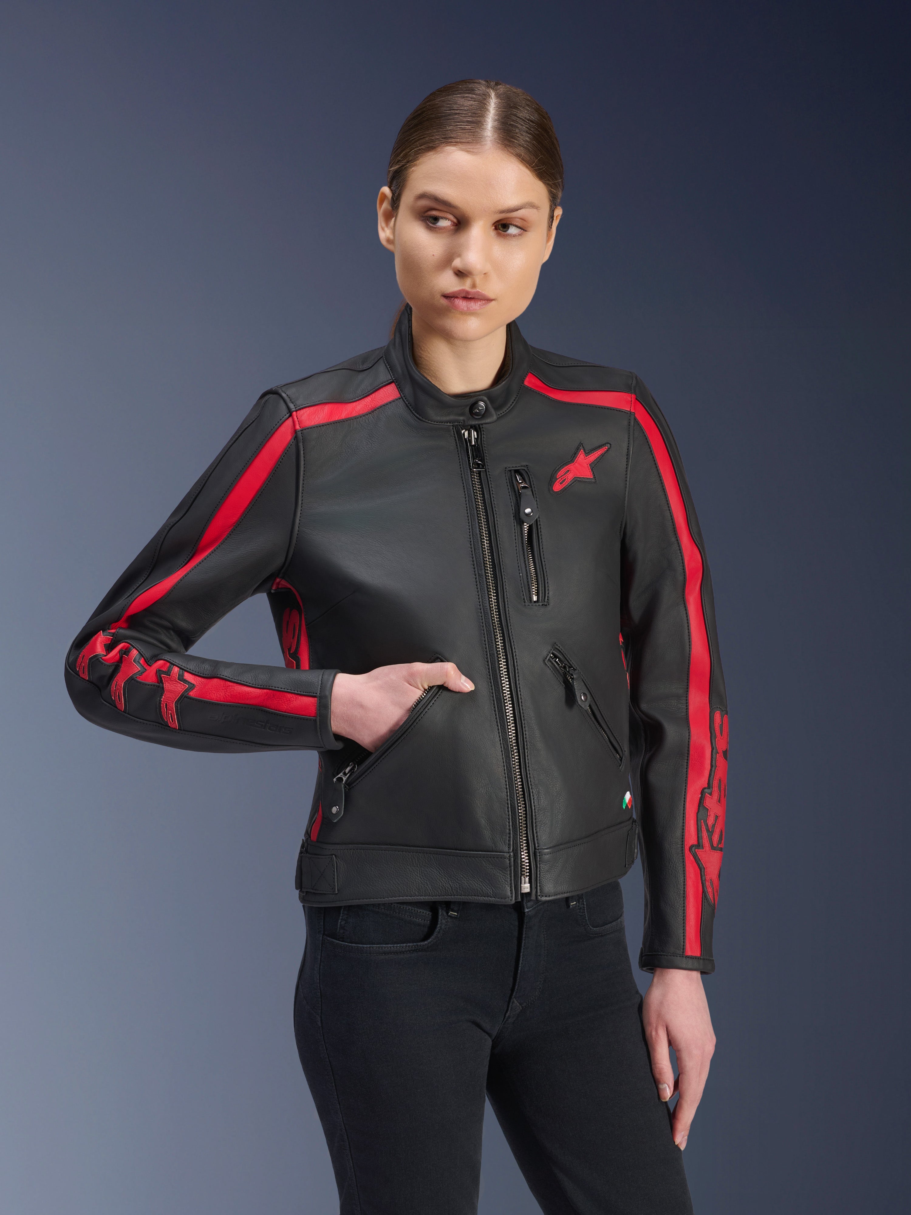 Giacca in pelle Alpinestars Stella Dyno, giacca da moto da donna, nero e rosso, indossata da una modella con jeans neri, con strisce racing rosse su spalle e maniche e branding evidente, realizzata in pelle premium, design d'ispirazione heritage per la guida urbana