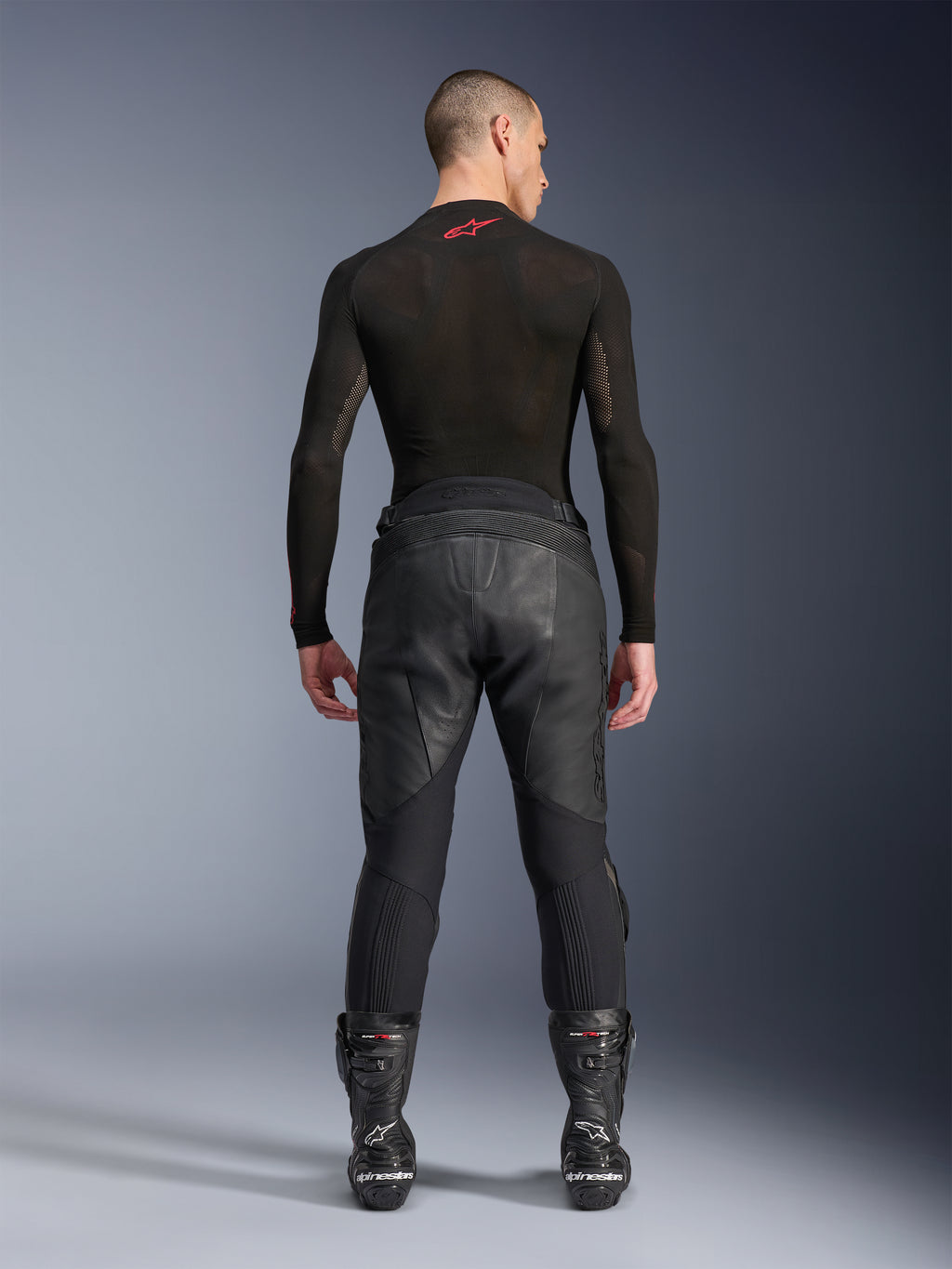 Pantaloni in pelle Alpinestars Missile V3, nero, vista posteriore su modello che indossa una maglia a compressione nera a maniche lunghe e stivali racing, costruzione in pelle premium con sovrapposizione a vita alta e zone elasticizzate, abbigliamento professionale da motociclismo
