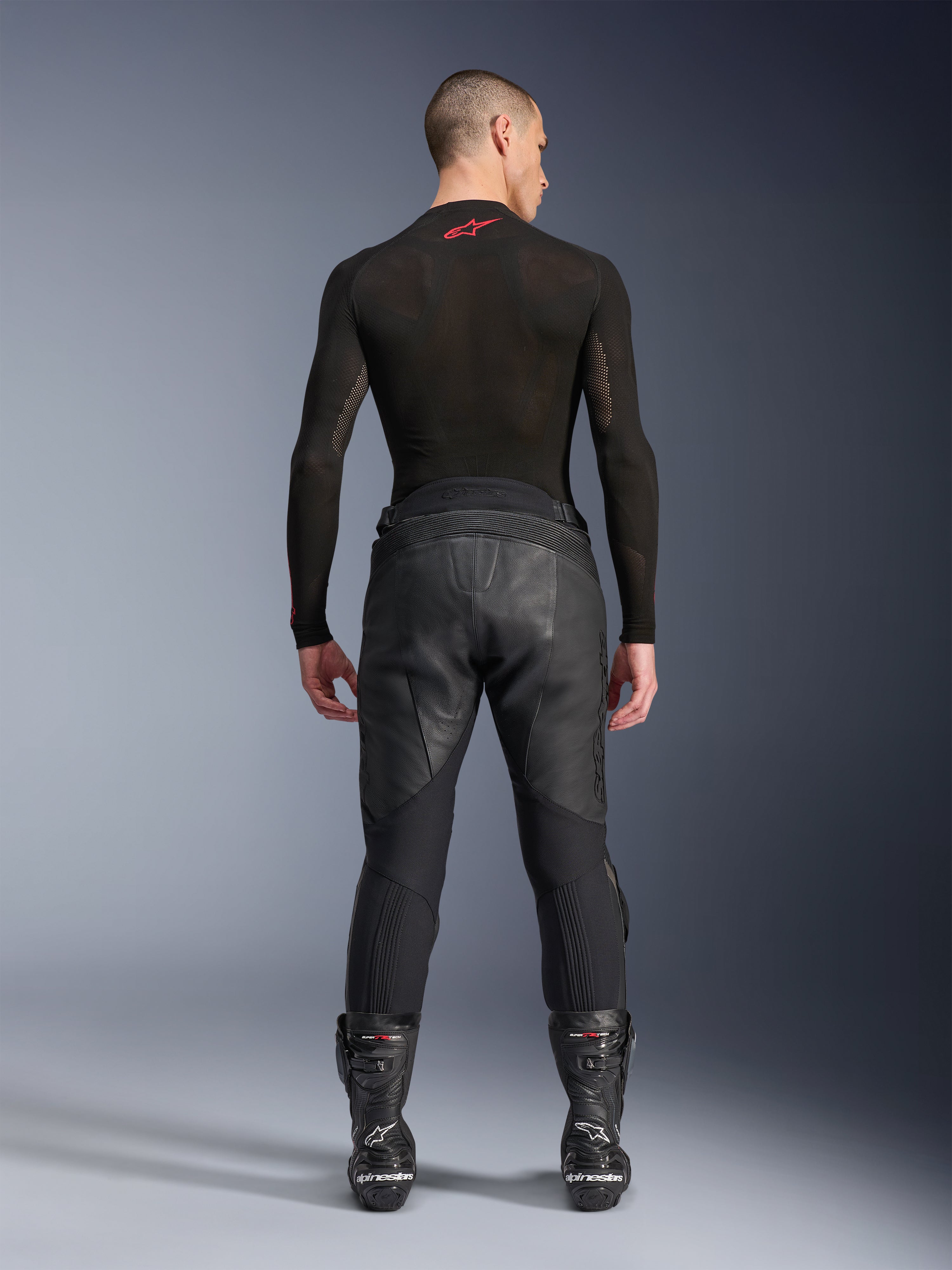 Pantaloni in pelle Alpinestars Missile V3, nero, vista posteriore su modello che indossa una maglia a compressione nera a maniche lunghe e stivali racing, costruzione in pelle premium con sovrapposizione a vita alta e zone elasticizzate, abbigliamento professionale da motociclismo