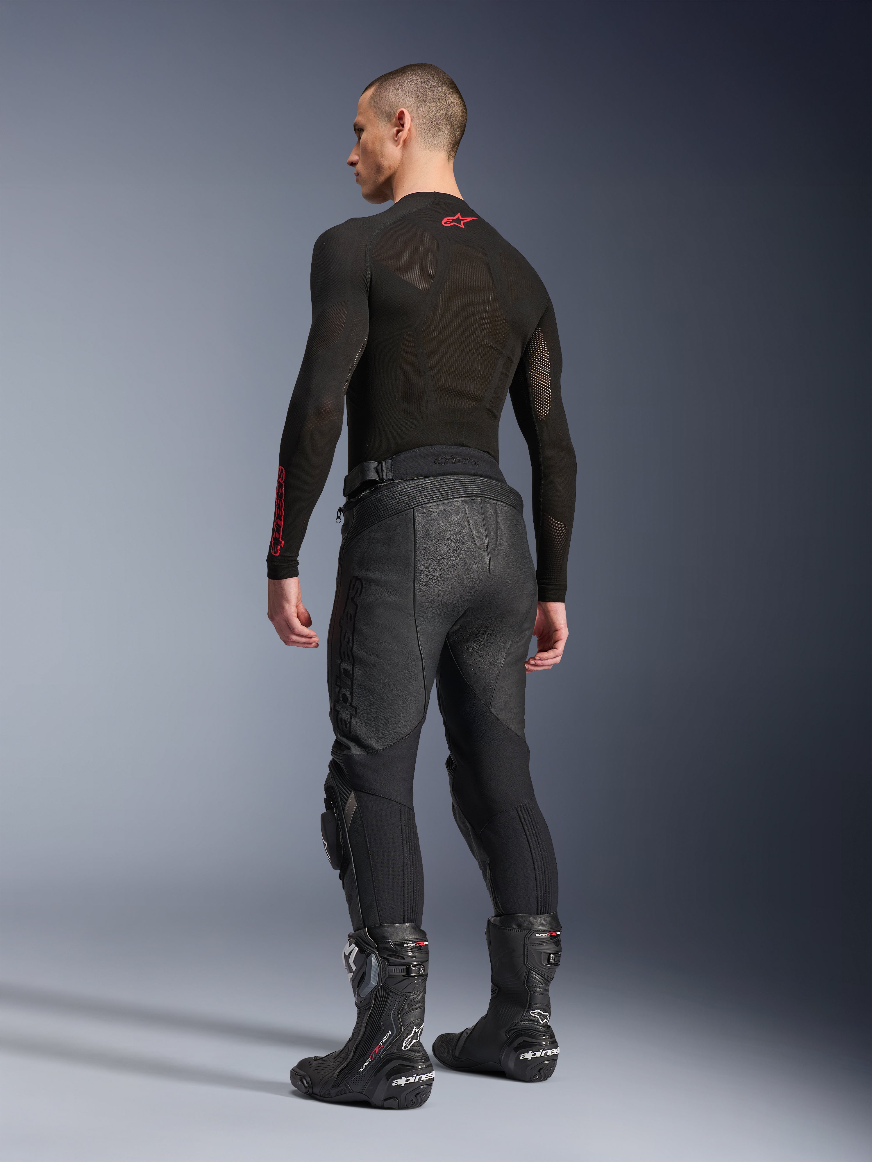 Pantaloni in pelle Alpinestars Missile V3, nero, vista posteriore su modello, indossati con un base layer nero a maniche lunghe e stivali da corsa motociclistici neri, caratterizzati da pannelli elastici in pelle a fisarmonica e un design pre-curvato per una vestibilità ergonomica durante la guida.