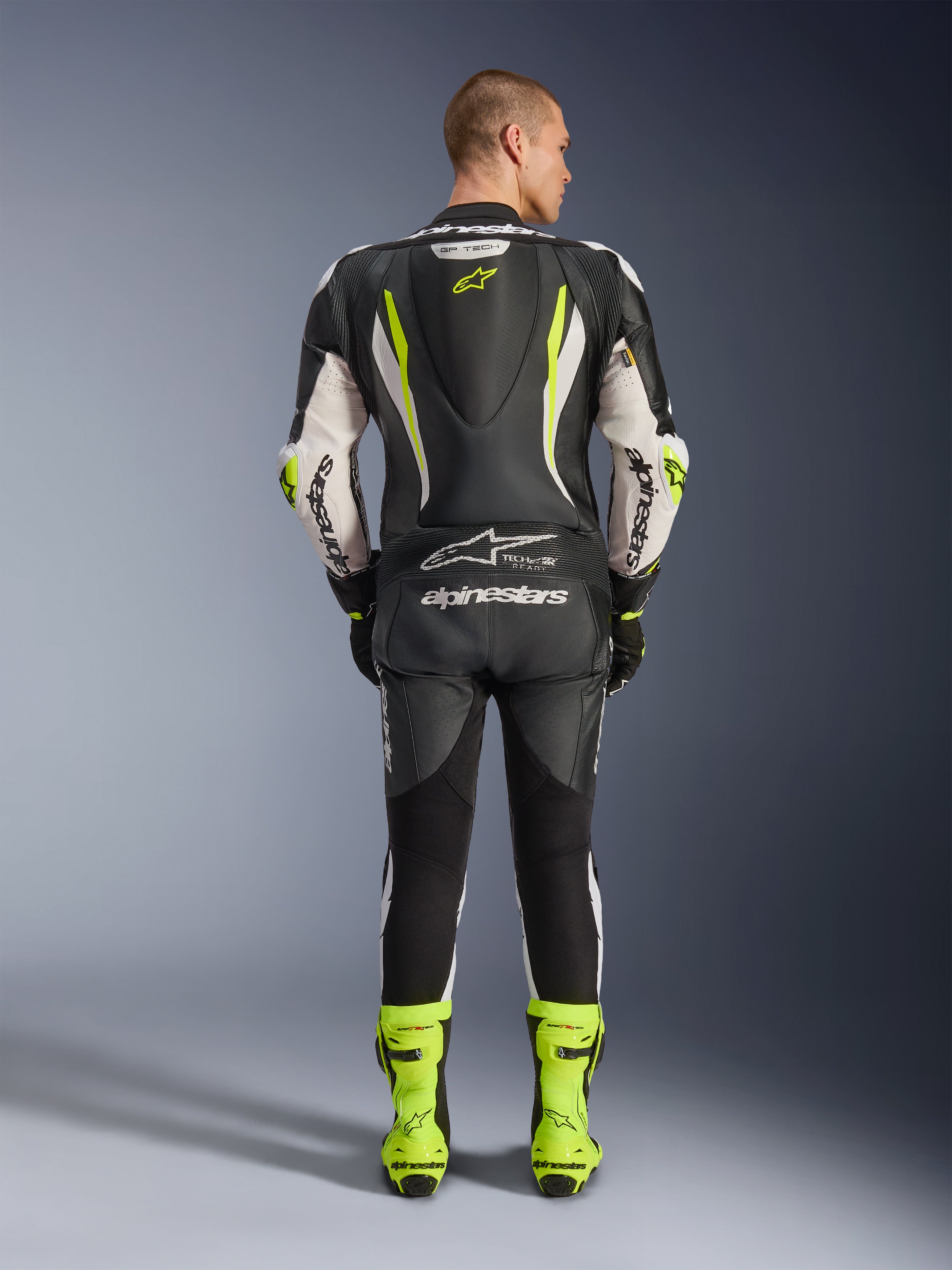 Alpinestars GP Tech V5 One Piece Leather Suit, vista posteriore, nero bianco e giallo fluo, con gobba aerodinamica e logo a stella giallo, marchio Alpinestars bianco su maniche e schiena, stivali da corsa coordinati in giallo neon, abbigliamento professionale da pista per moto