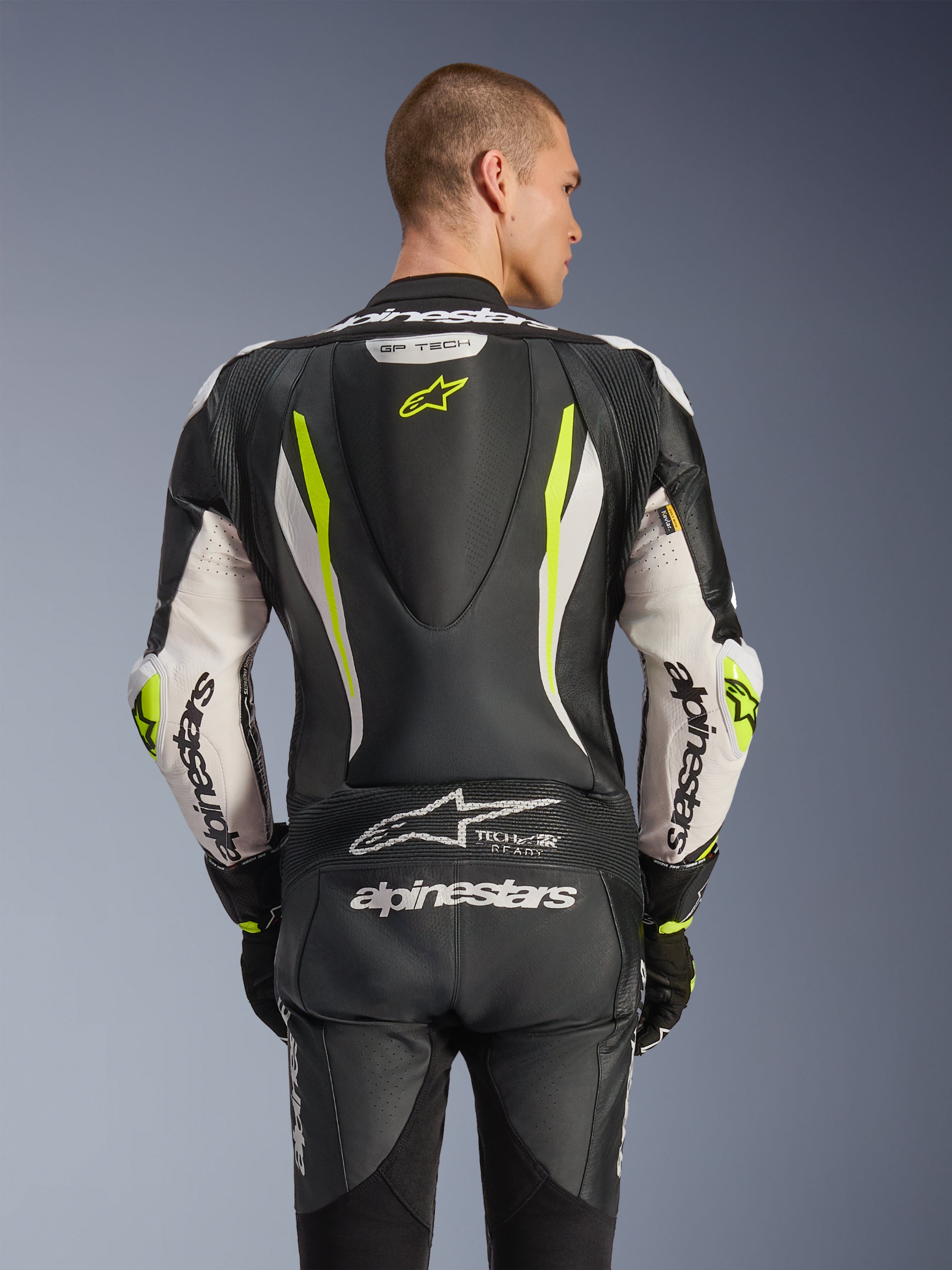 Alpinestars GP Tech V5 One Piece Leather Suit, vista posteriore su modello, nero, bianco e giallo fluo, con gobba aerodinamica, grandi loghi Alpinestars sulle braccia e sulla seduta, pannelli elasticizzati a fisarmonica e logo Tech-Air Ready