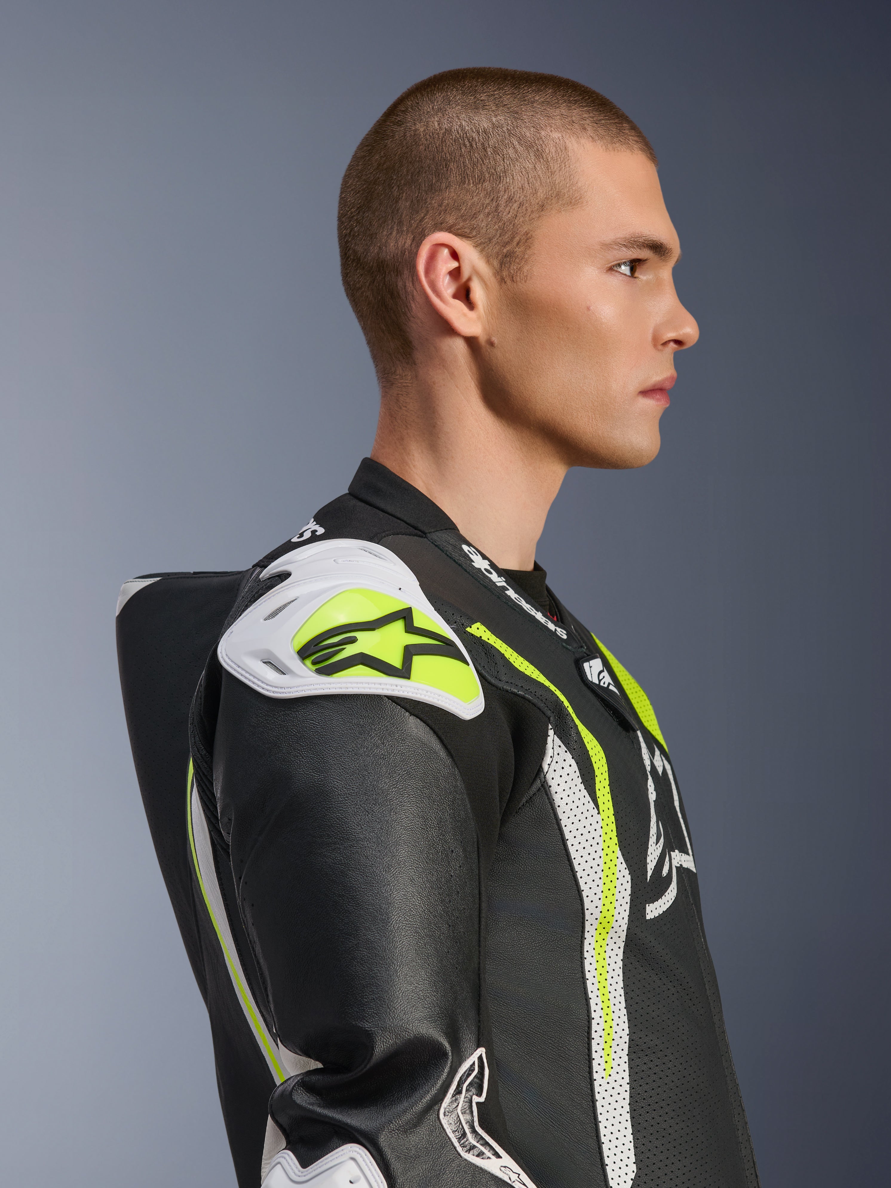 Modello che indossa la tuta in pelle intera Alpinestars GP Tech V5 in nero bianco giallo fluo, vista laterale di profilo che evidenzia la gobba aerodinamica e gli slider bianchi sulle spalle, abbigliamento protettivo da moto di ispirazione racing con dettagli in pelle traforata